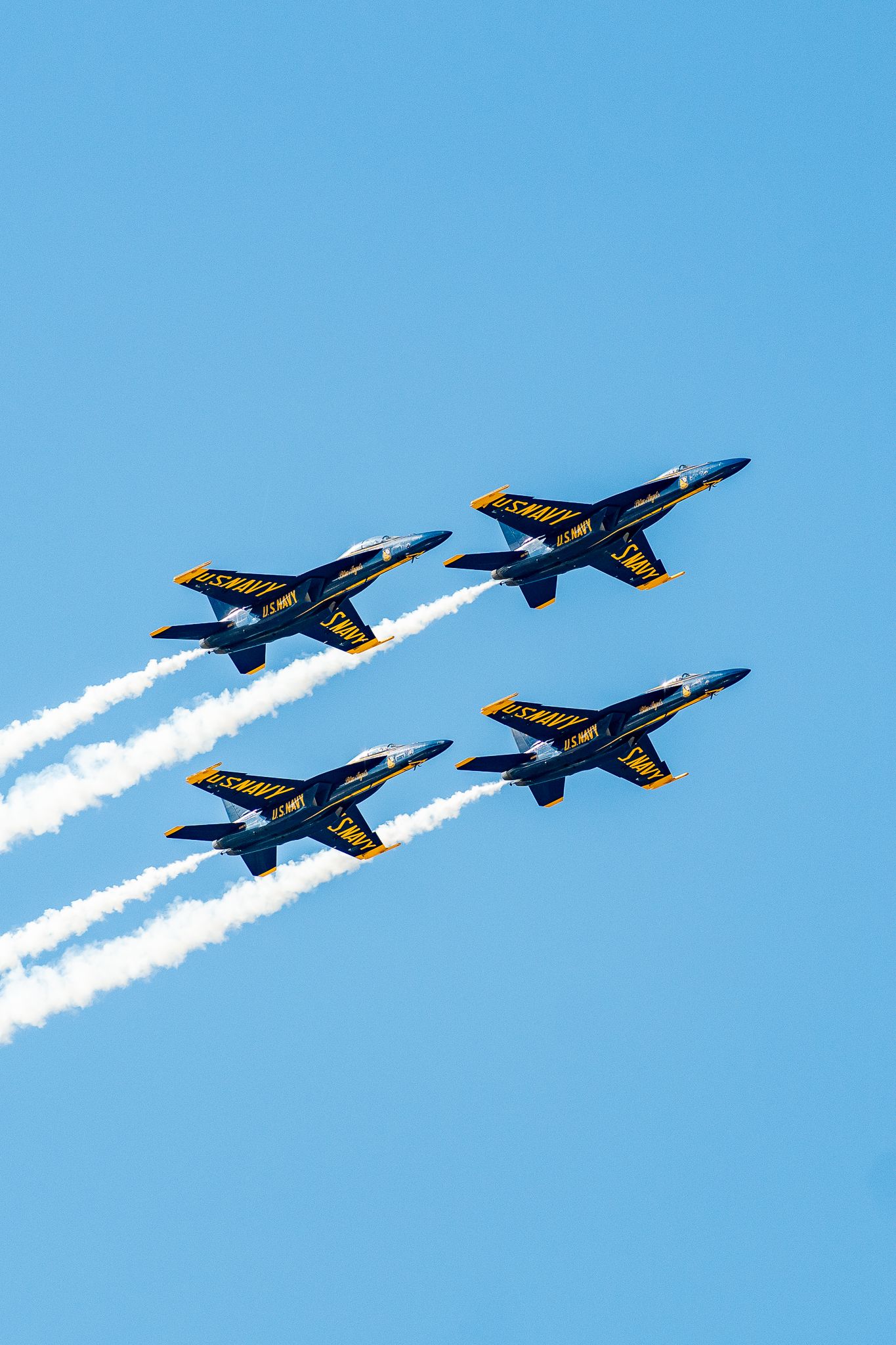 US Navy Blue Angels flying in diamond formation with white smoke trails against a clear blue sky.