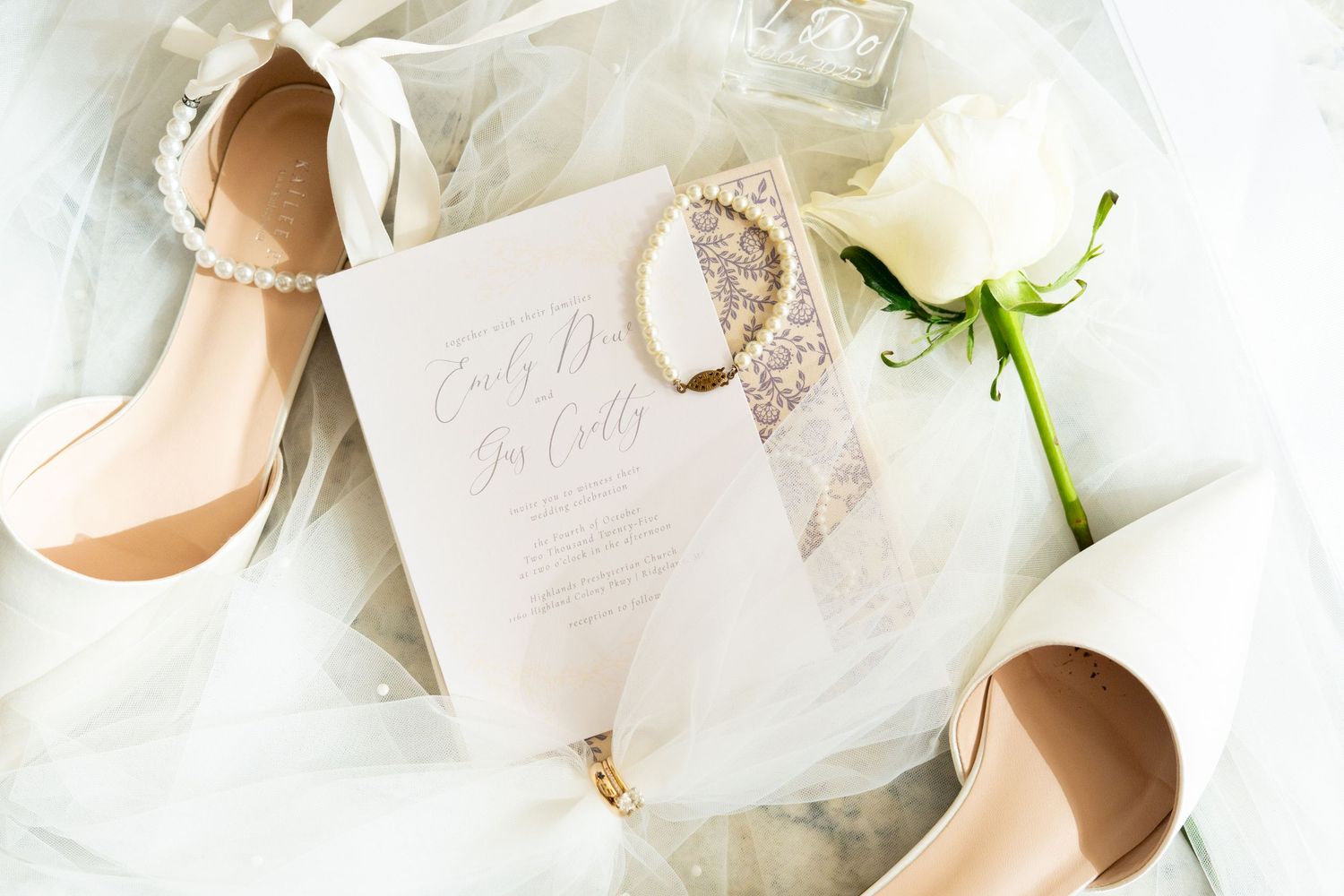 Elegant bridal flat lay with nude heels, pearl bracelet, white rose, and wedding invitation on ivory tulle.