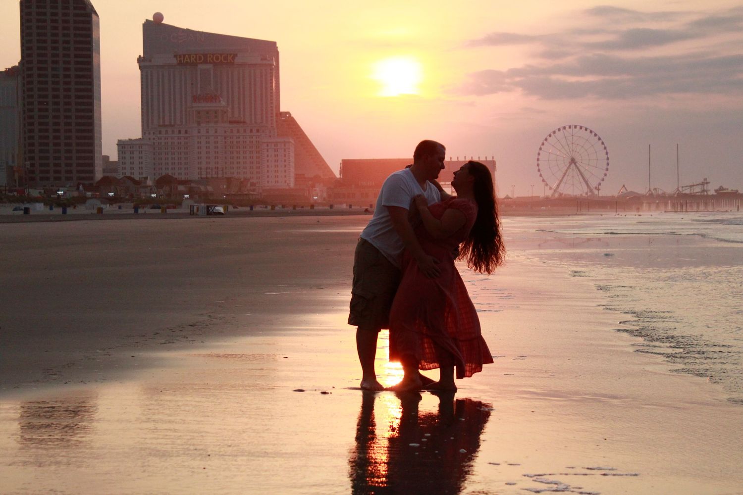 A silhouetted couple embraces on a beach at sunset with a ferris wheel in the background.