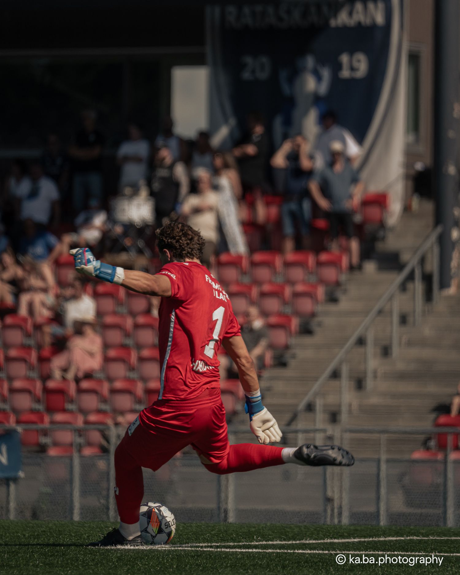 Goalkeeper in red uniform kicking the ball during a soccer match in a stadium with red seats.