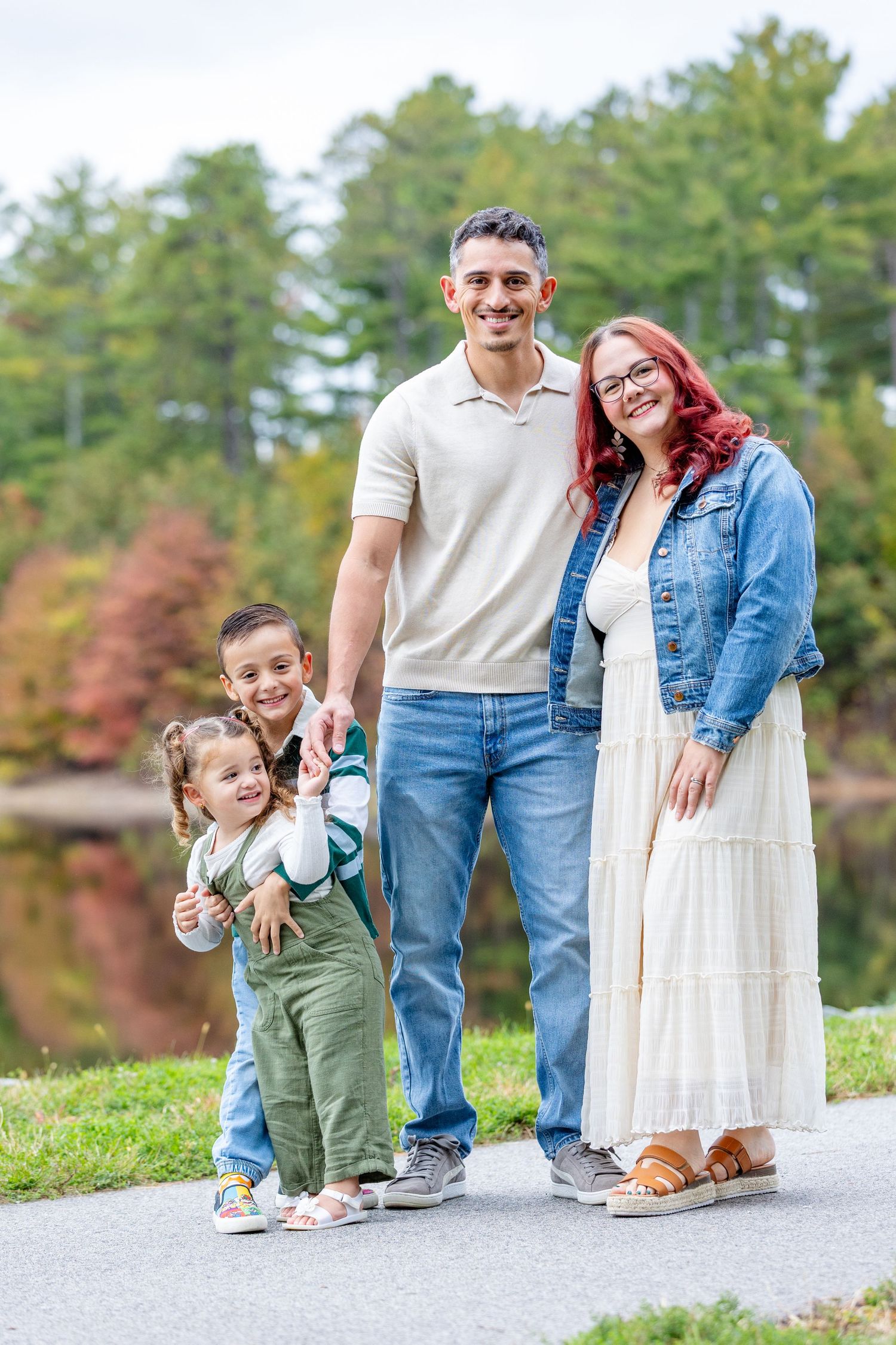 Family wearing casual clothing stands together outdoors on a paved path with autumn trees in background.
