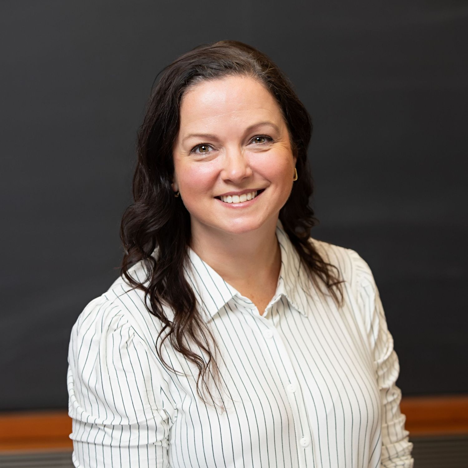 Professional headshot of a smiling woman with dark hair wearing a white striped shirt against a dark background.