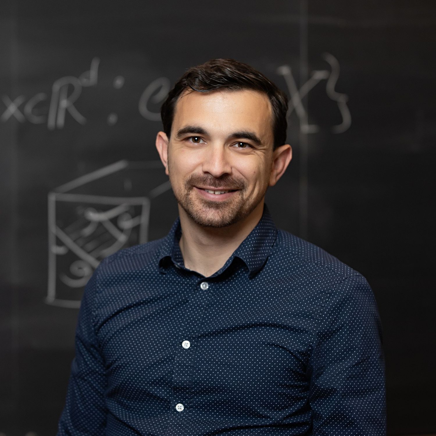 A smiling man in a dark blue dotted shirt stands confidently in front of a chalkboard with mathematical equations.