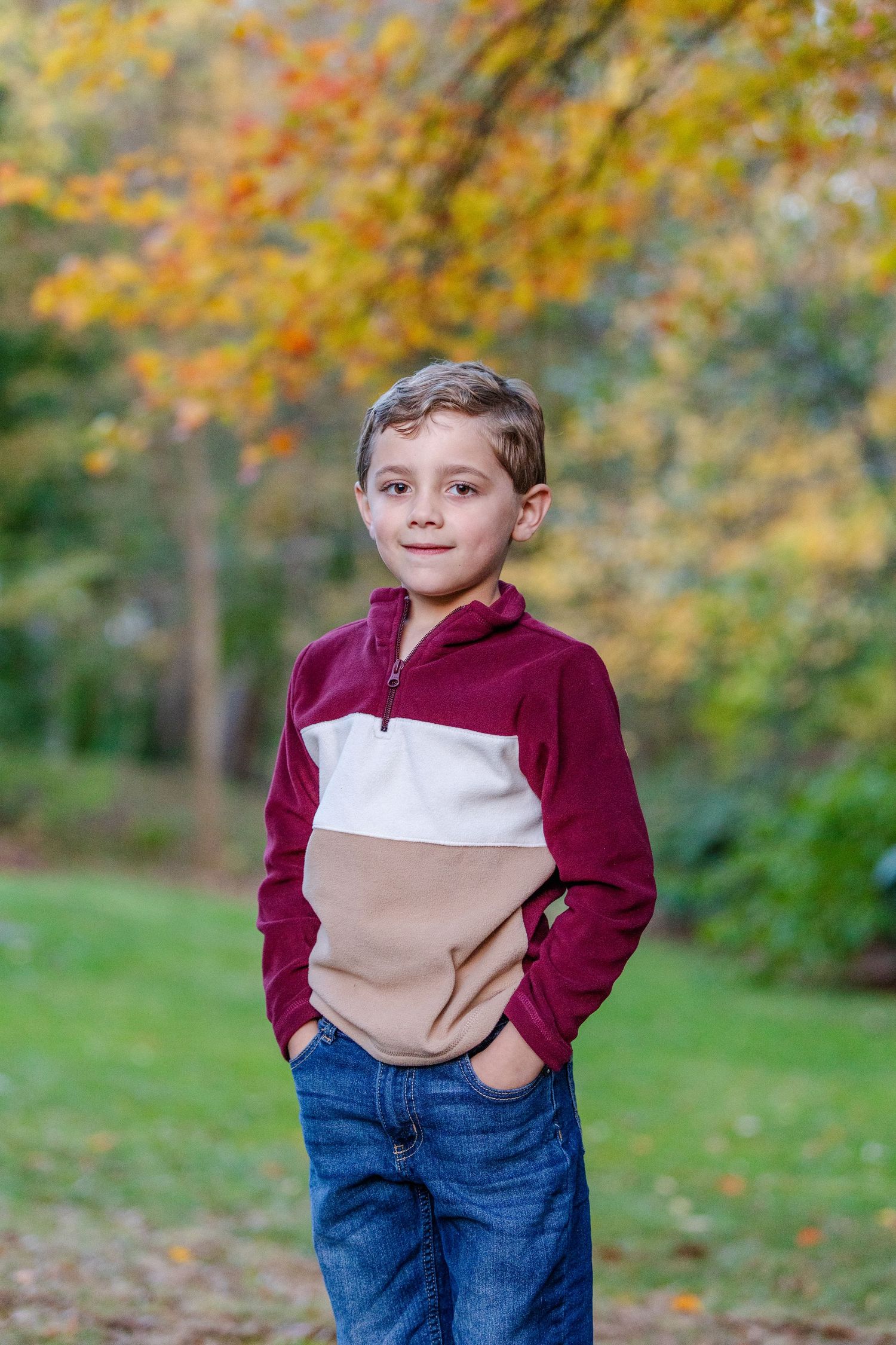 A young person in a burgundy and tan color-block sweatshirt and jeans stands outdoors among autumn foliage.