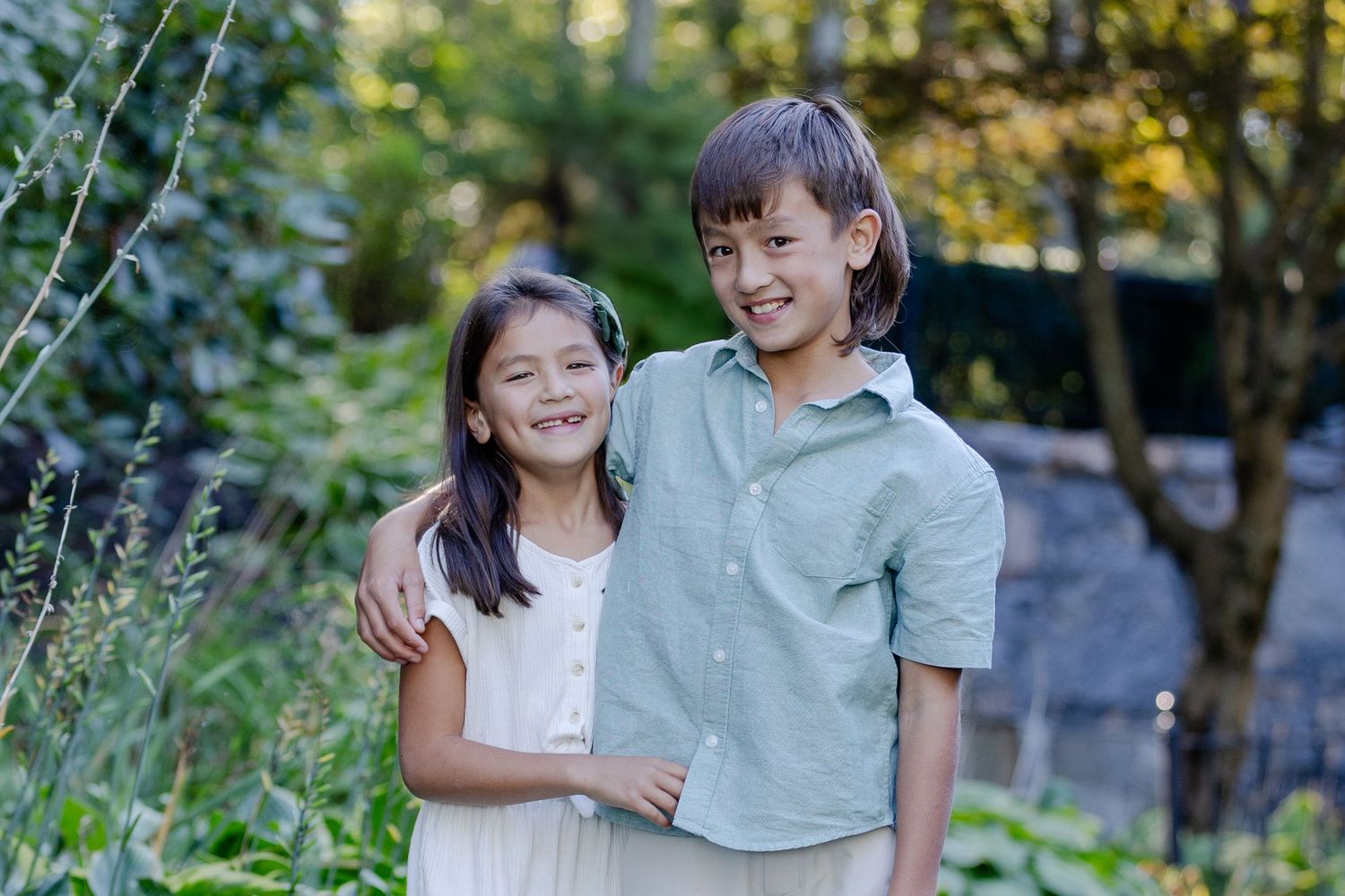 Two smiling children posing together in a lush green garden, wearing casual summer clothes.