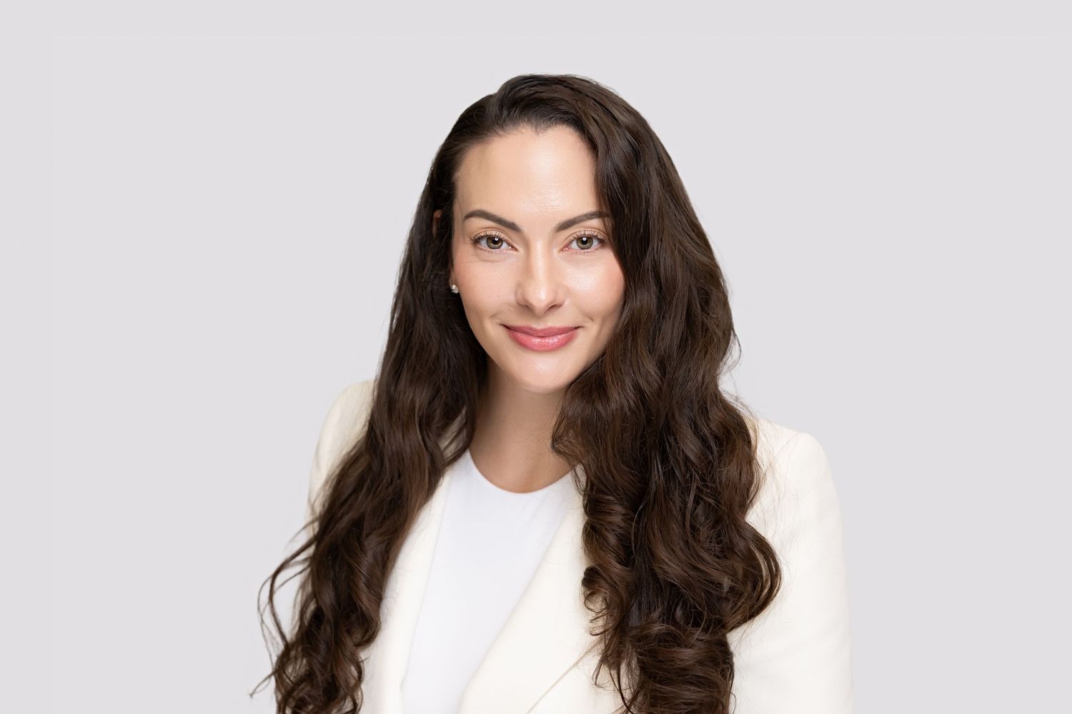 Professional headshot of a young woman with long wavy brown hair, wearing a white blazer, smiling against a gray background.