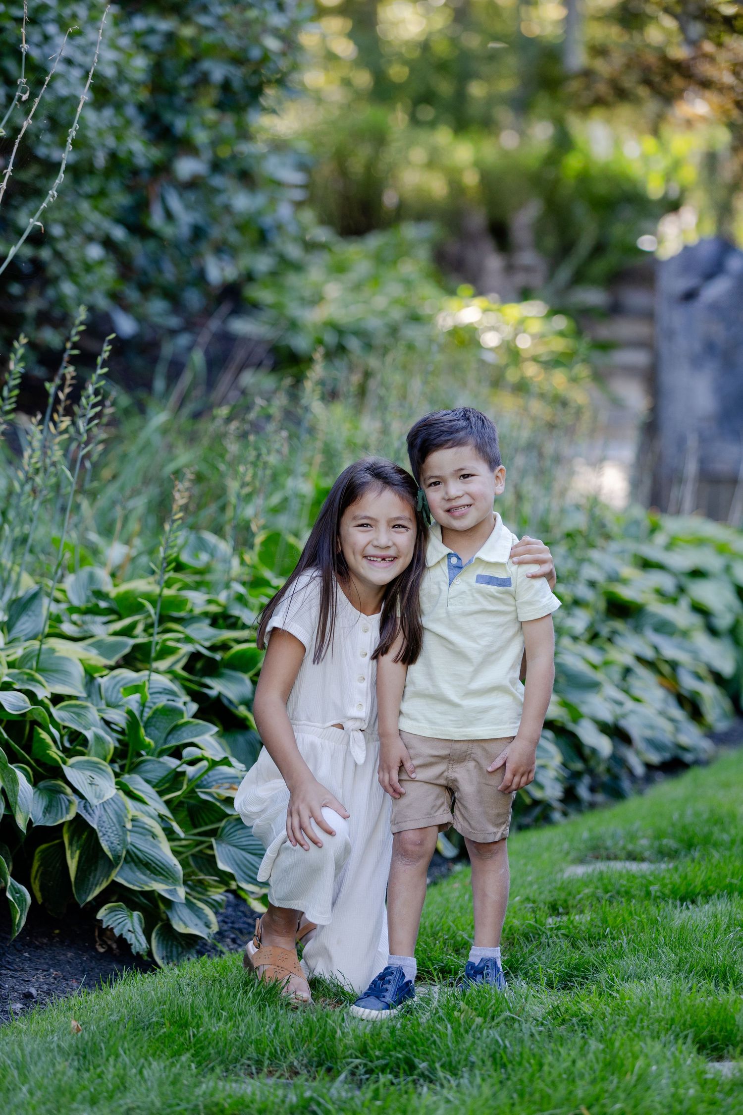 Two children sit together and laugh while playing outdoors in a lush garden setting.