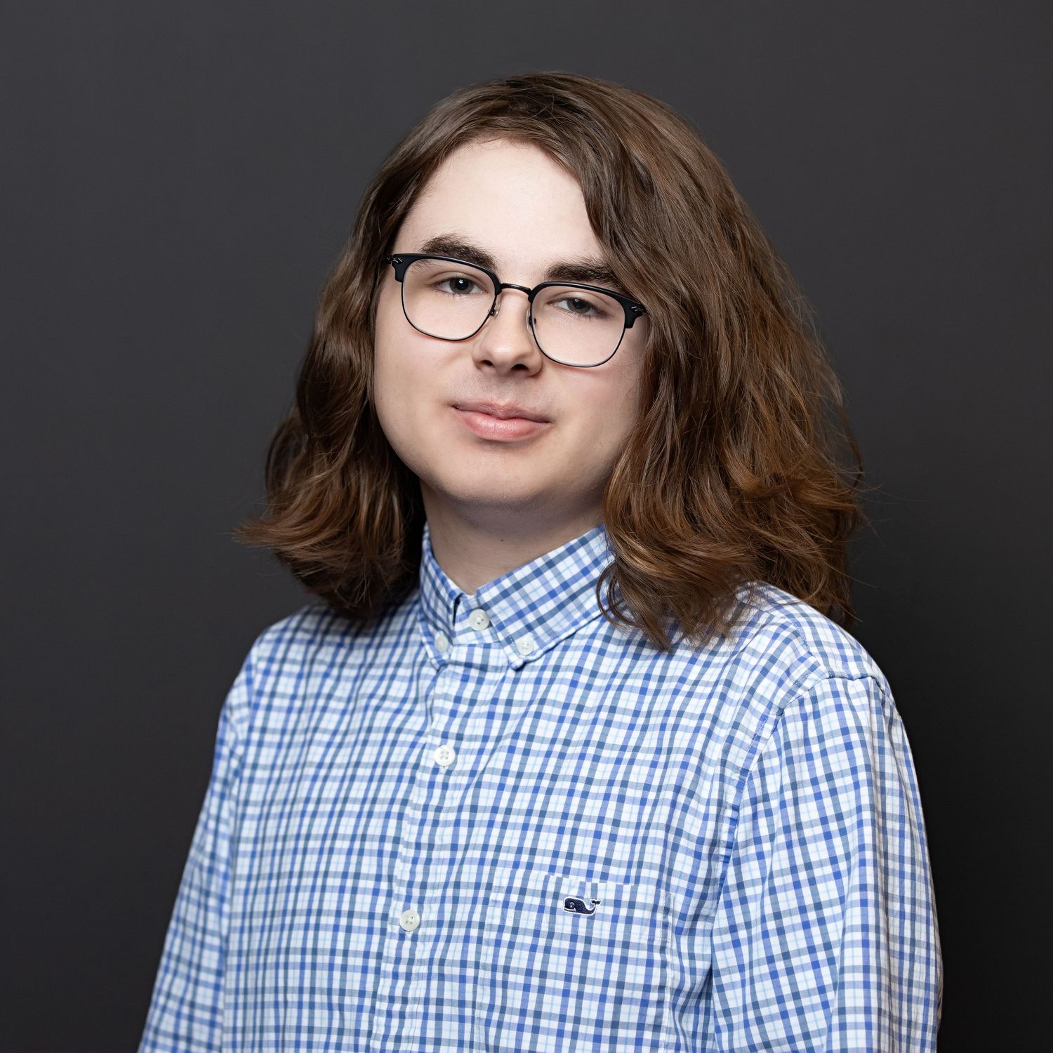 Young person with shoulder-length brown hair and glasses wearing a blue plaid shirt against a dark gray background.