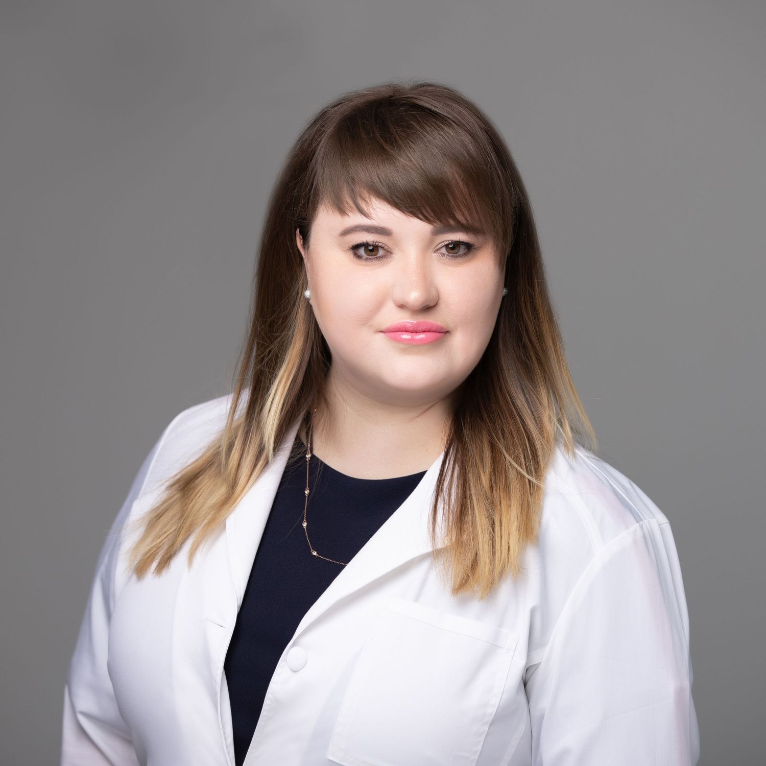 Professional headshot of a young woman in a white lab coat and dark top, with ombre hair, against a gray background.