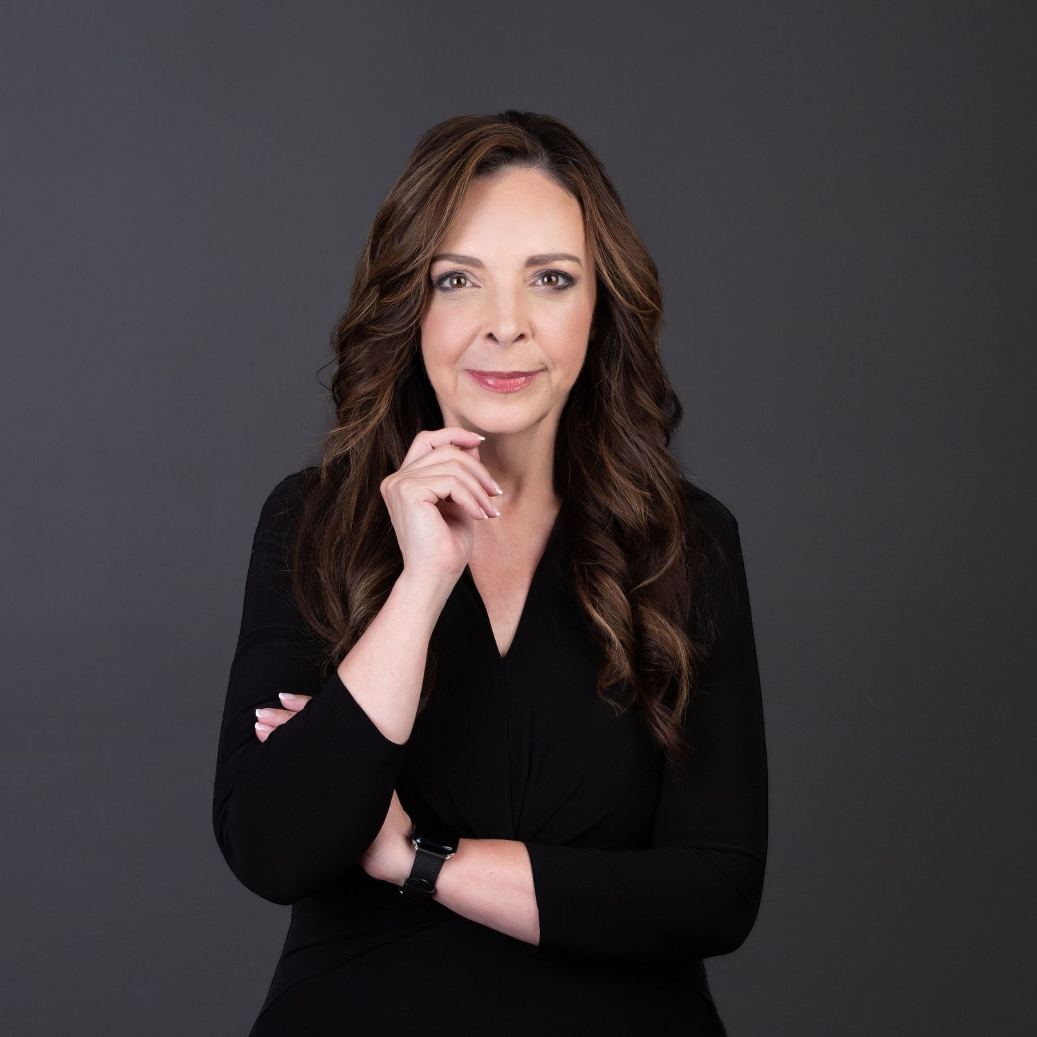 Professional woman in black blazer poses confidently against gray background with hand on chin.