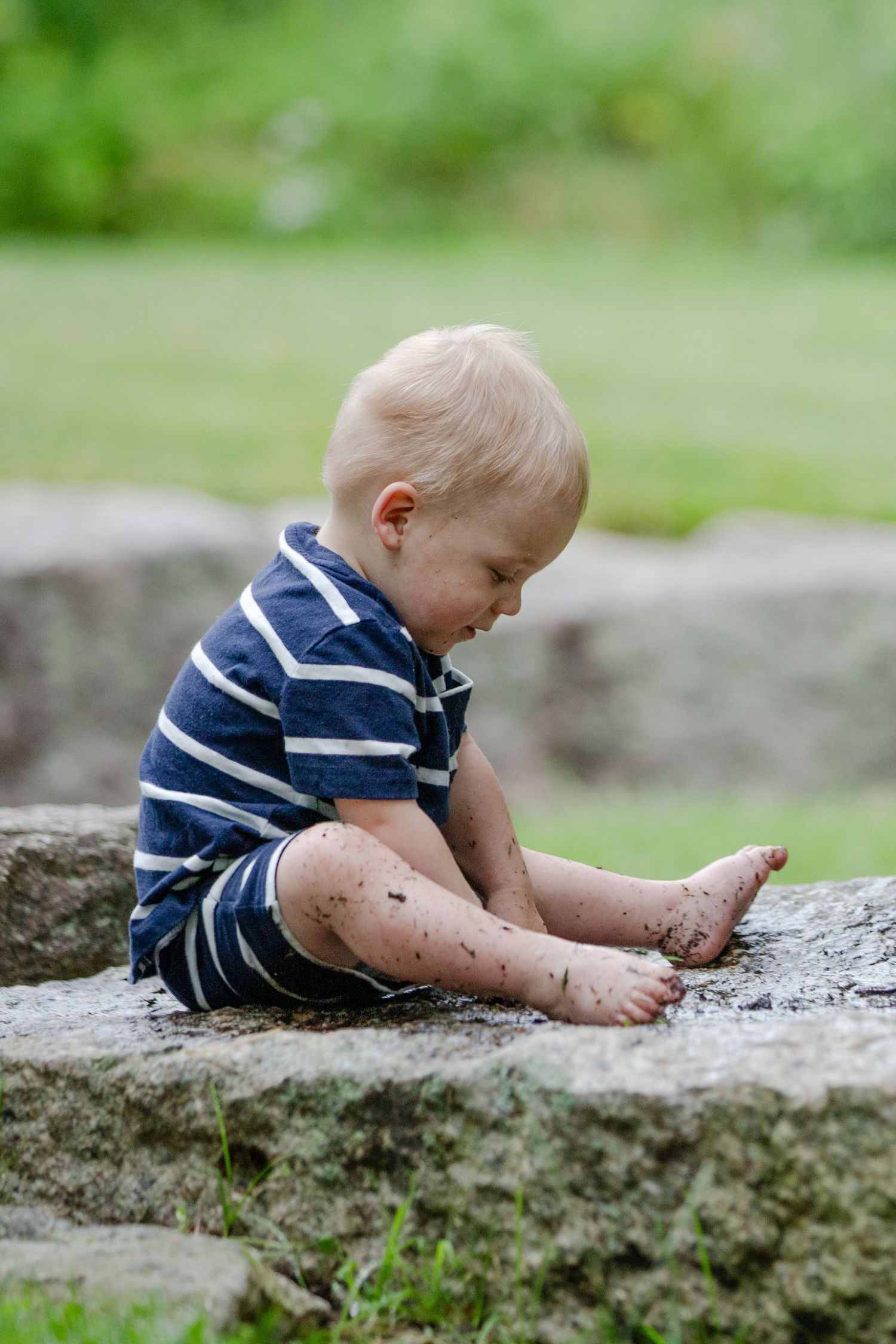 A small child in striped shirt plays and explores in mud on a stone wall on a sunny summer day.