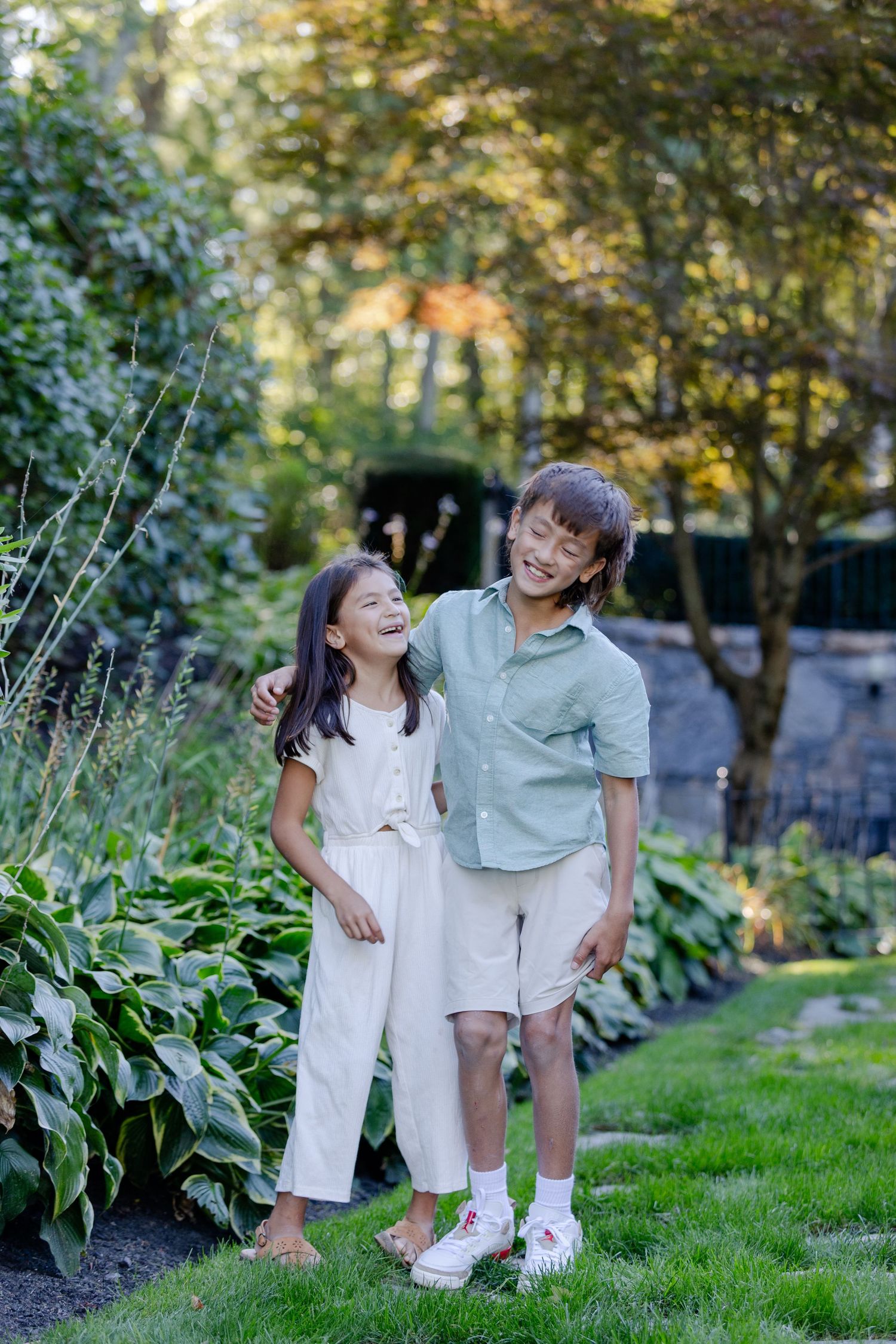 Two happy children laughing and embracing in a lush green garden, wearing white and sage green outfits.