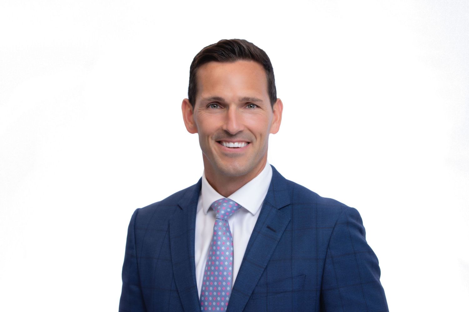 Professional headshot of a smiling man in a navy blue suit and lavender tie against a white background.