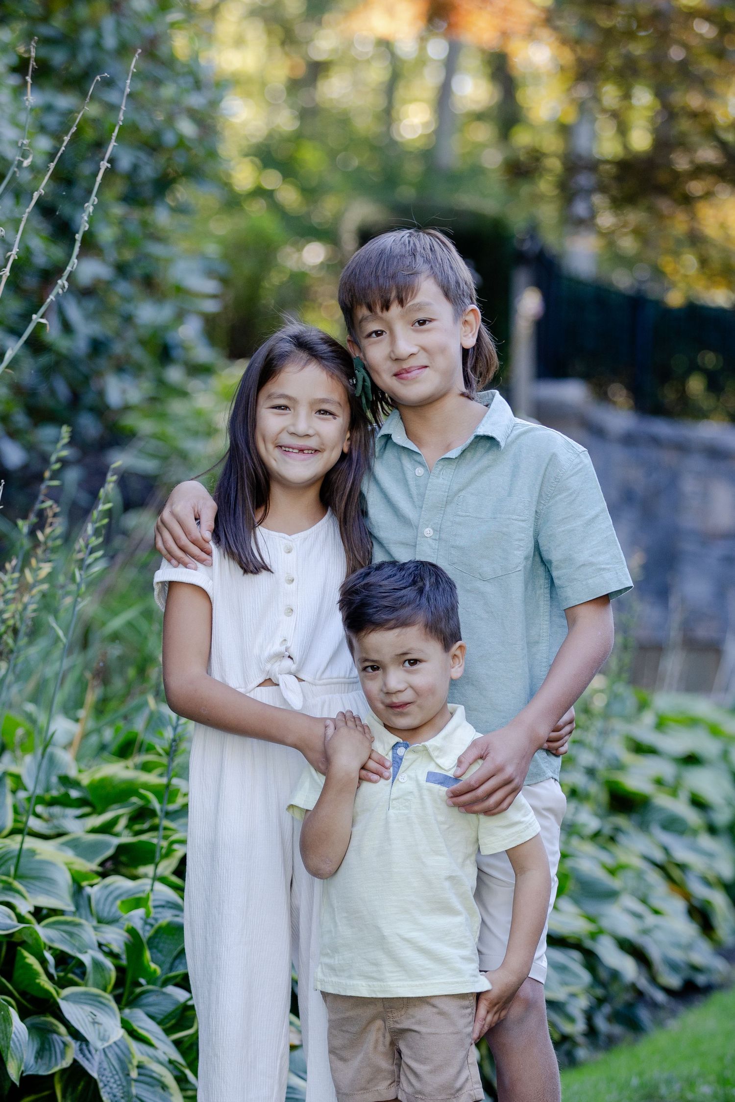 Three children stand together in a garden setting with lush greenery and natural lighting in the background.
