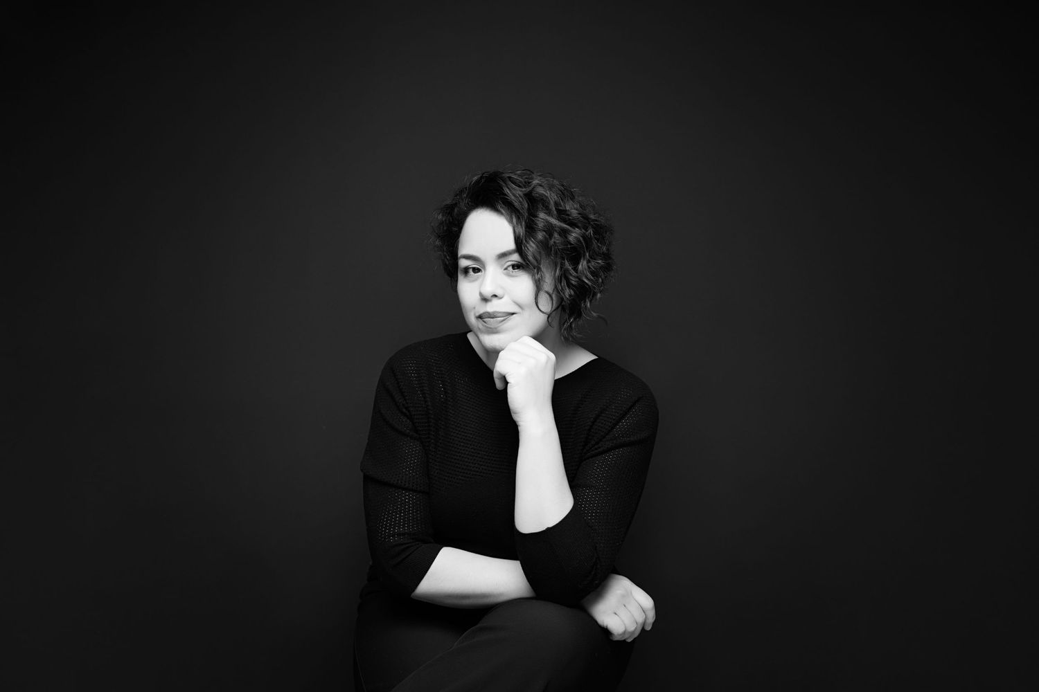 Black and white portrait of a woman in black top, sitting thoughtfully with hand on chin against dark background.