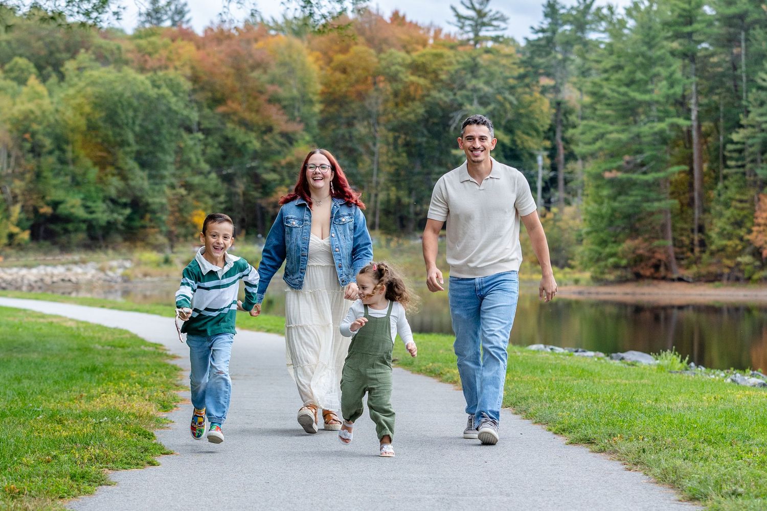 Family walking together on grassy path near lake during autumn season with colorful fall foliage in background.