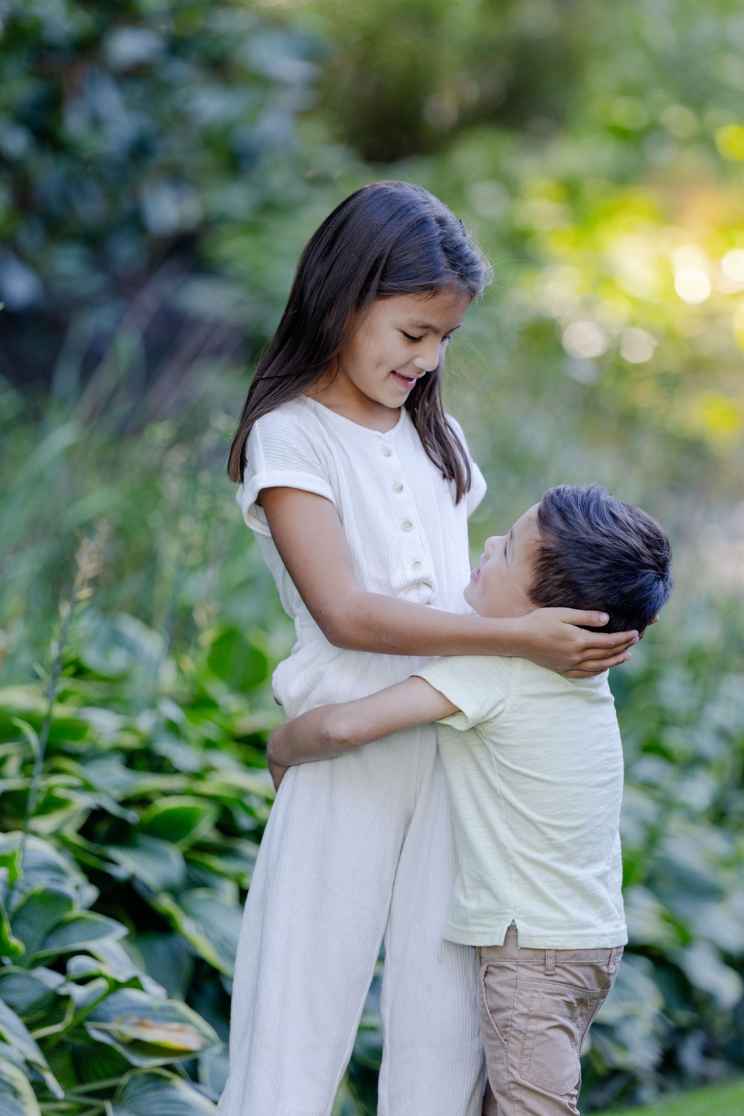 Two children in casual clothing share a tender embrace outdoors surrounded by lush green foliage.