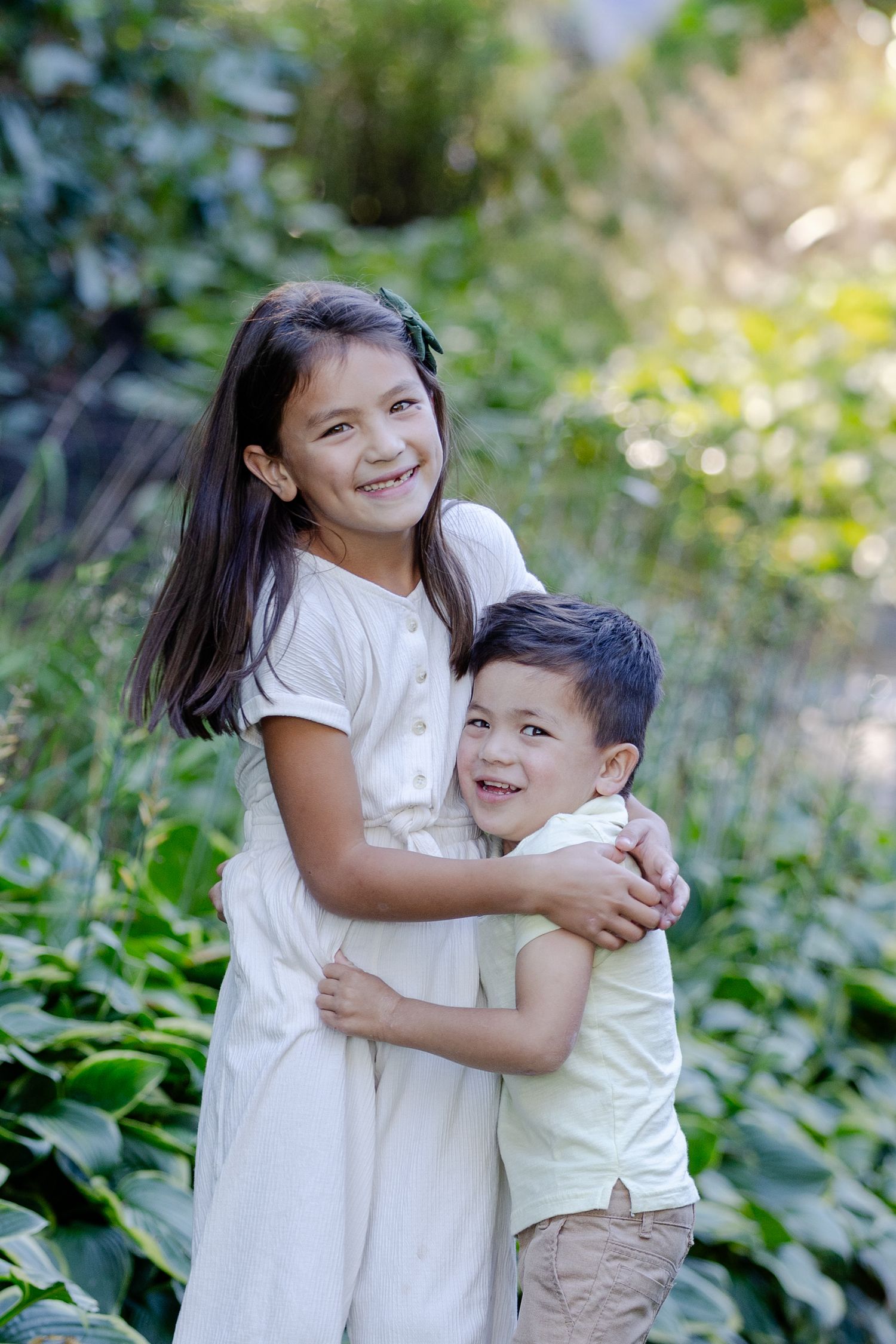 Two young children in white outfits share a joyful embrace while playing outdoors among green foliage.