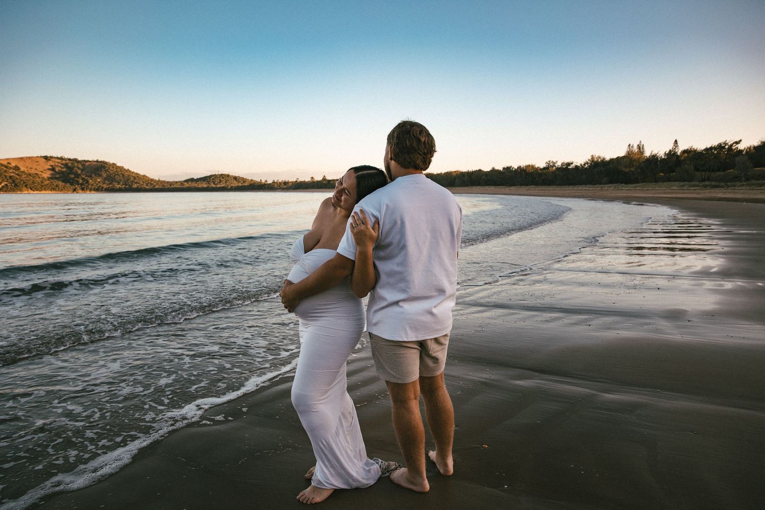 Couple embracing on a sandy beach at sunset, wearing white outfits, with calm ocean waves and hills in background.