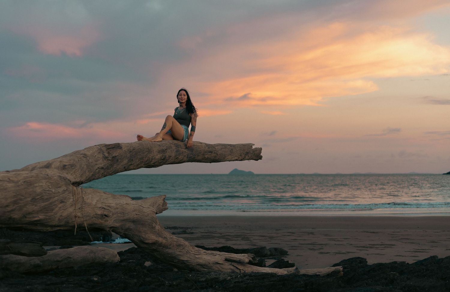 Woman sitting on driftwood at a tropical beach during a stunning pink and orange sunset with ocean views.