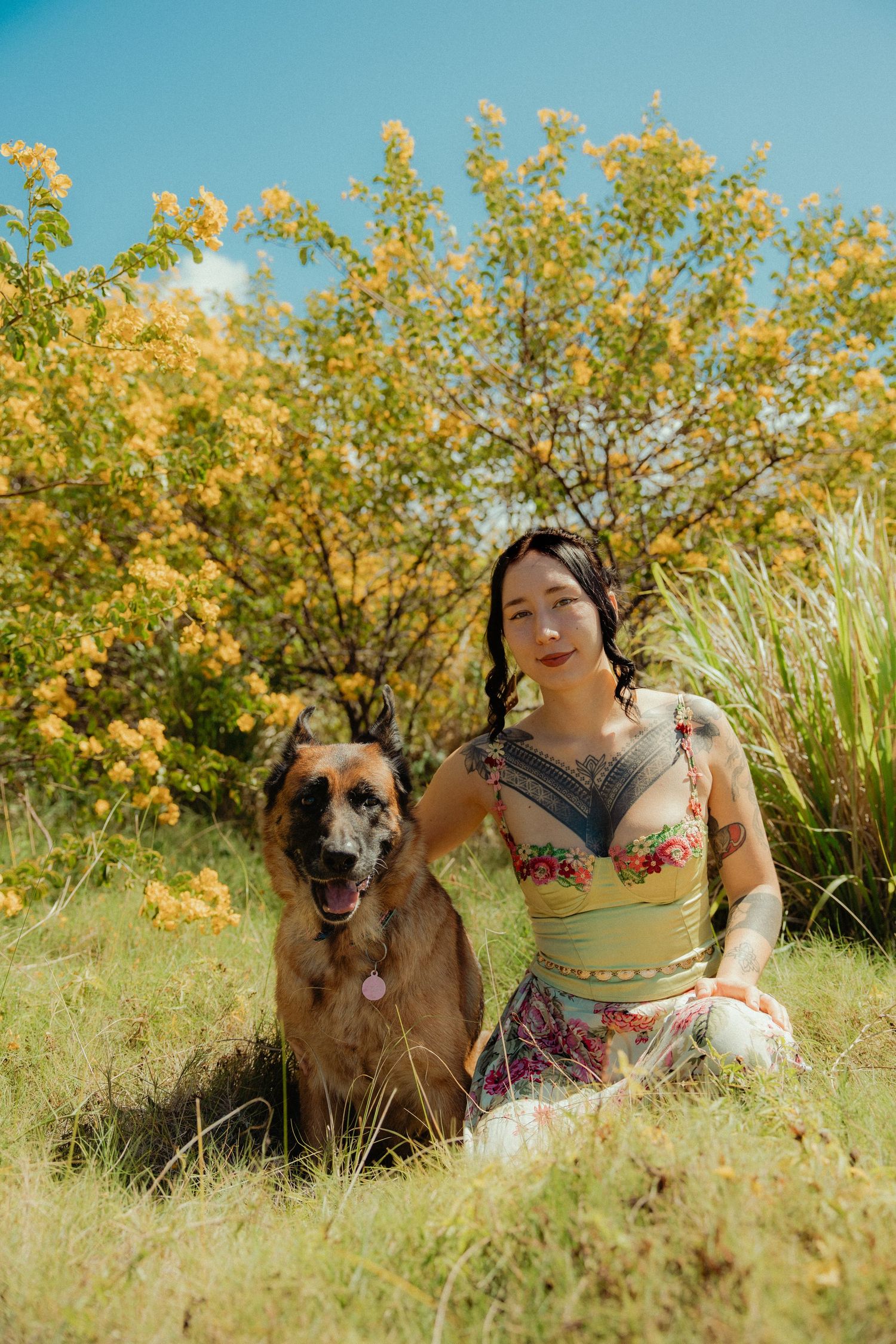 Woman with tattoos sits in green grass beside a German Shepherd dog, with yellow flowering bushes and blue sky behind them.