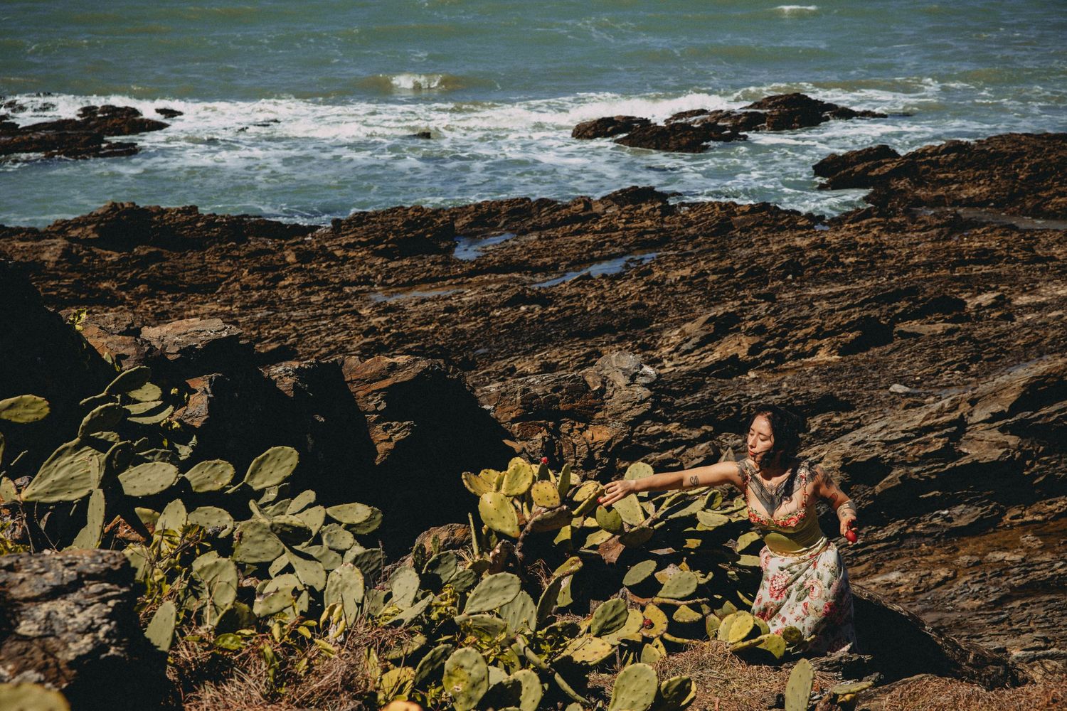 Woman sitting among tropical plants on rocky coastline with ocean waves and rugged rocks in background.