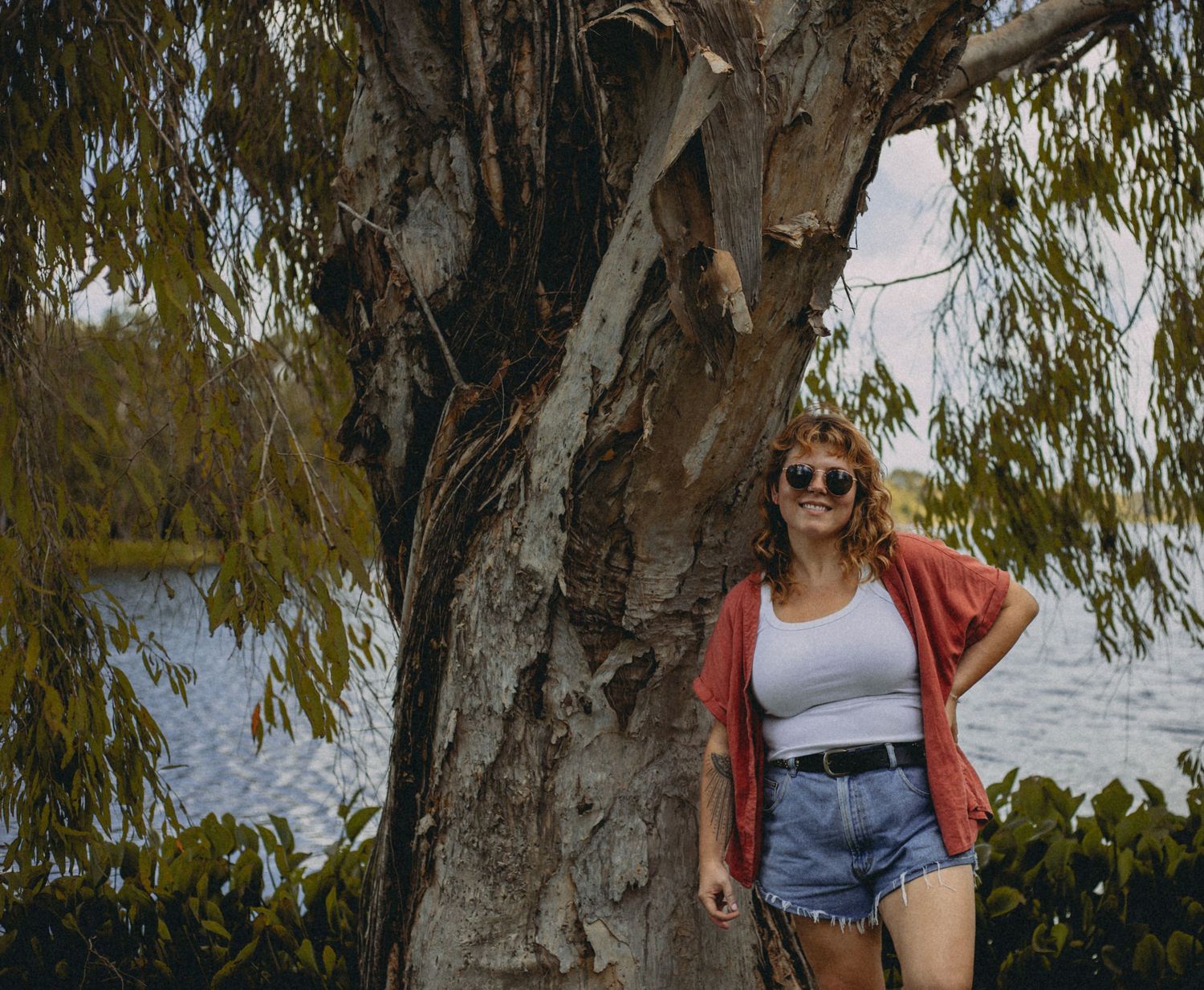 Young woman in sunglasses, white tank, denim shorts, and red cardigan leaning against a large tree near water.