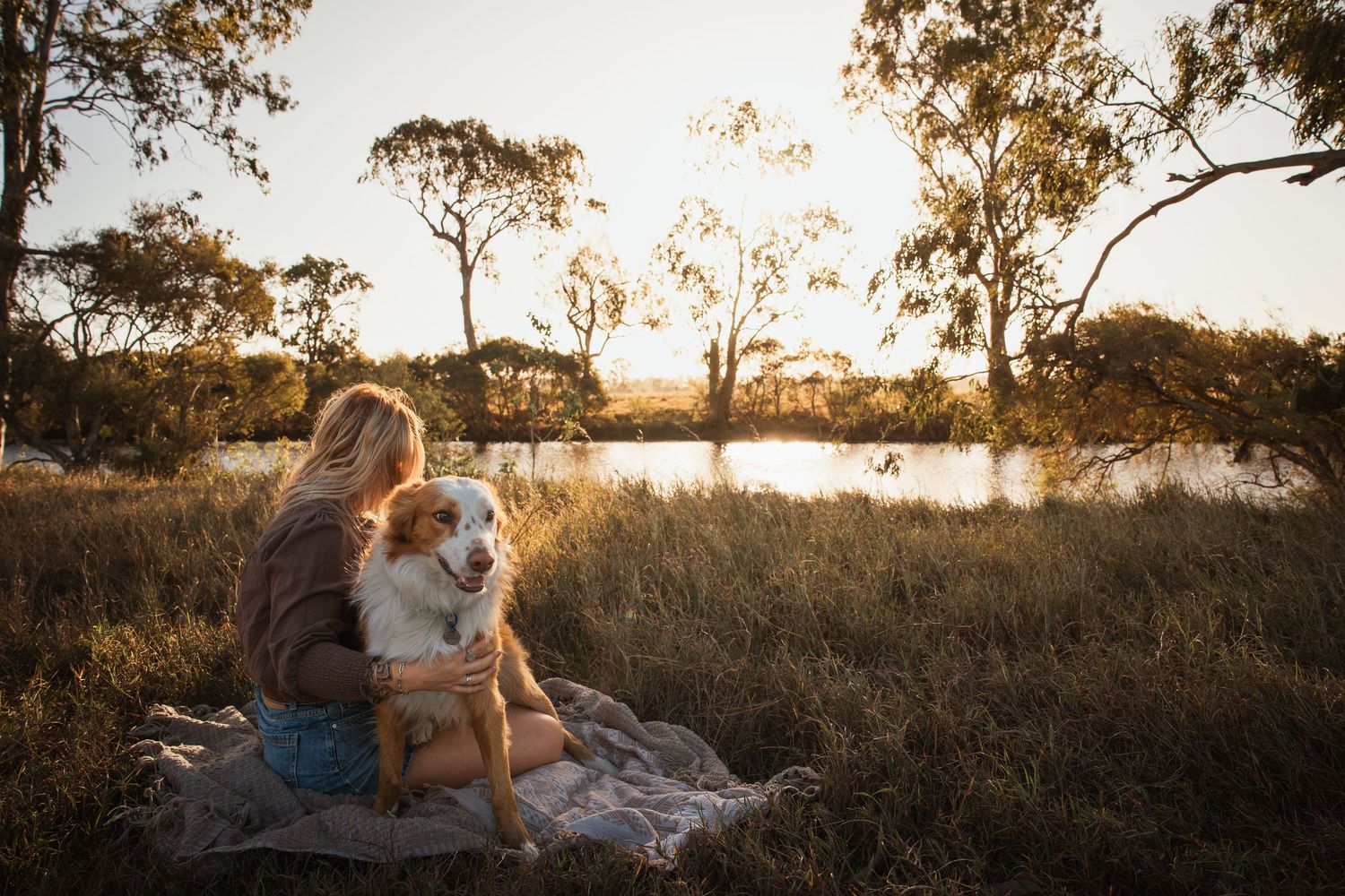 Woman sitting with Australian Shepherd dog on blanket in golden hour light near tranquil lake surrounded by eucalyptus trees.