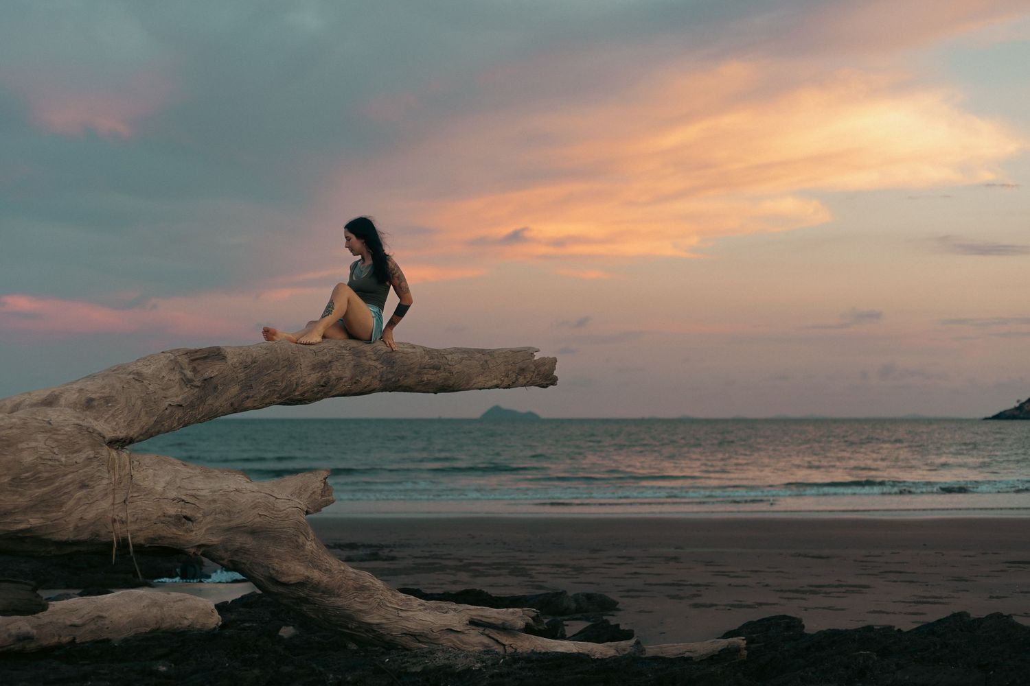 Woman in bikini sitting on driftwood at a tropical beach during a stunning pink and orange sunset.