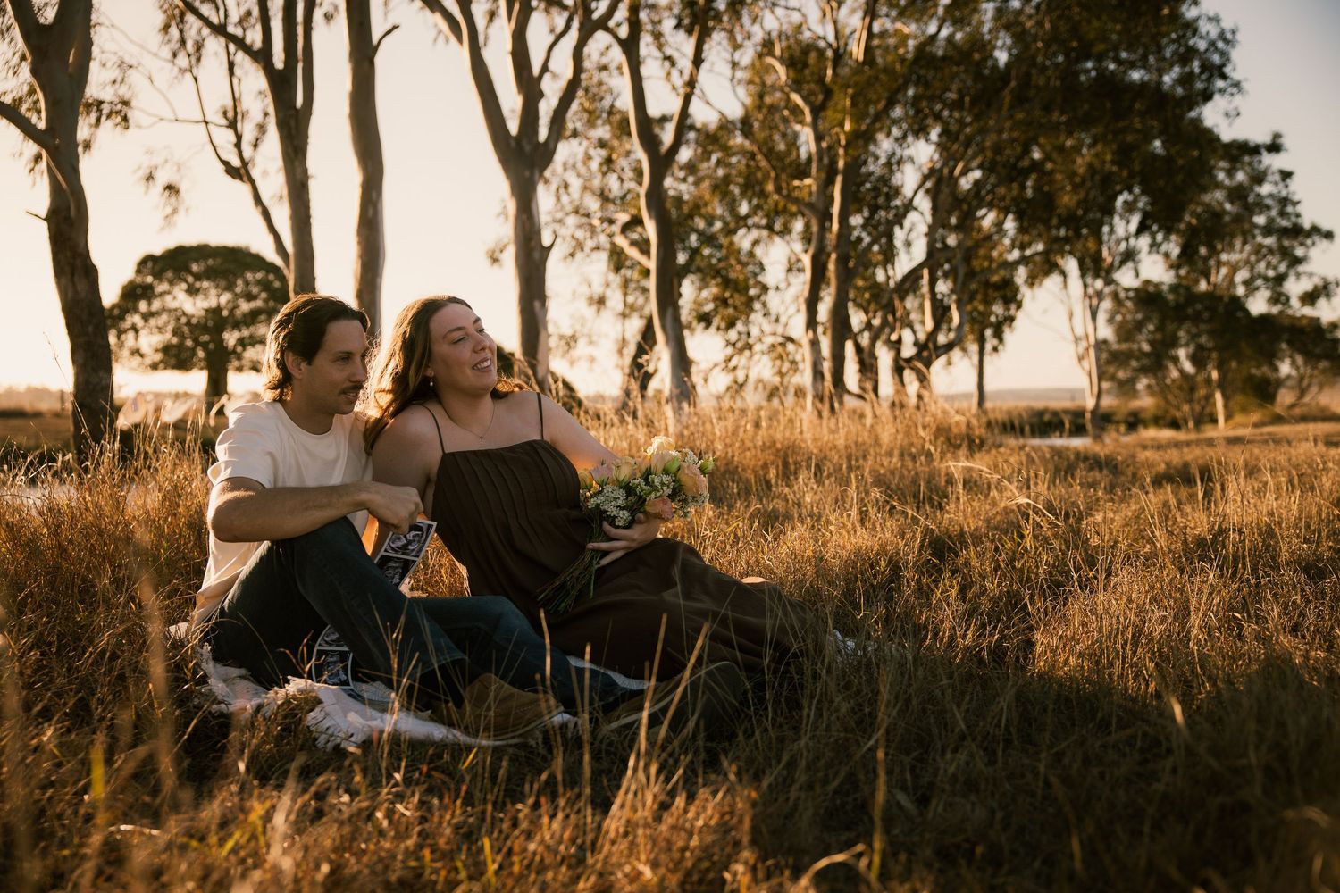 Couple sitting together in golden grass field at sunset, surrounded by tall trees in warm autumn light.