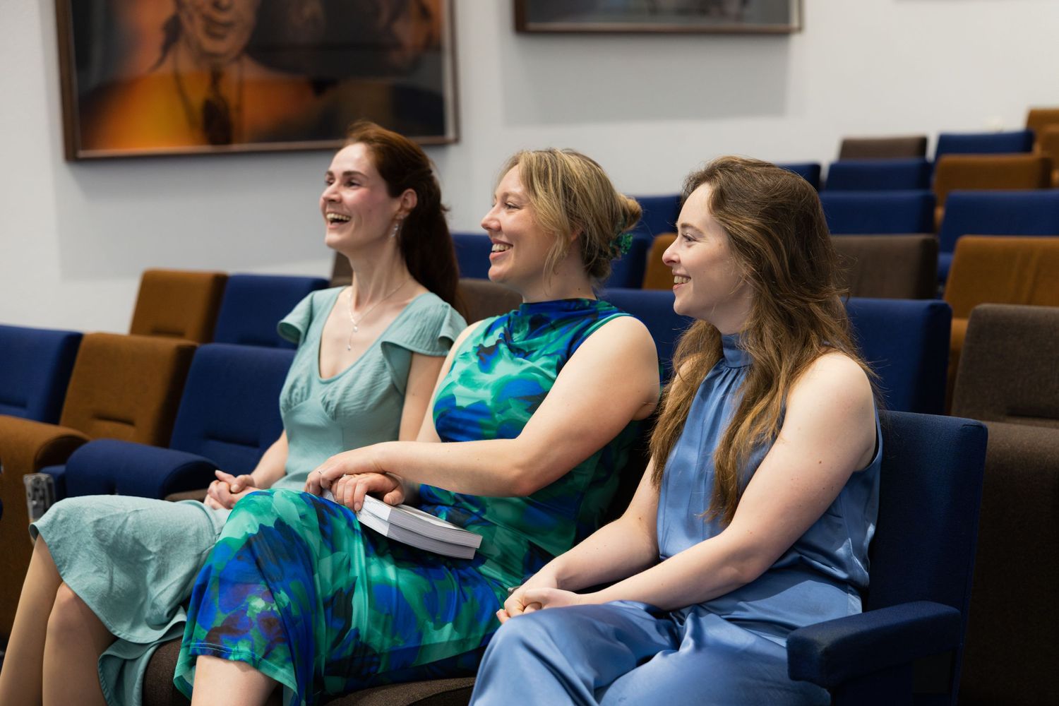 Three women in colorful dresses sit together smiling and laughing in blue auditorium chairs.