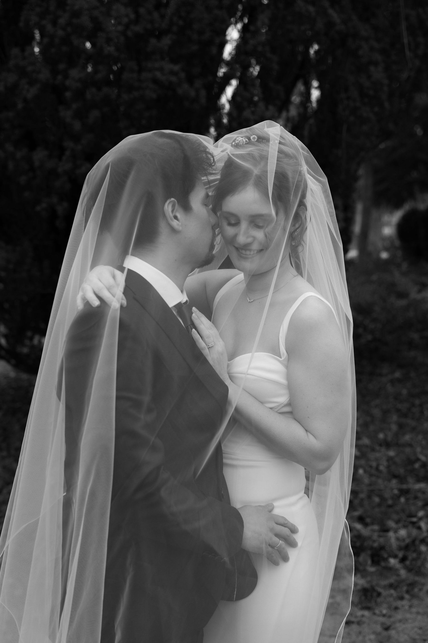 Black and white photo of bride and groom sharing romantic moment under wedding veil outdoors.