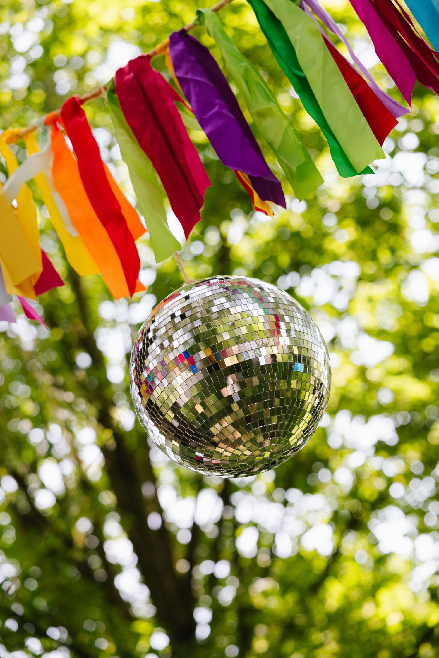 Shiny disco ball hanging outdoors under colorful rainbow ribbon bunting with green trees in the background.