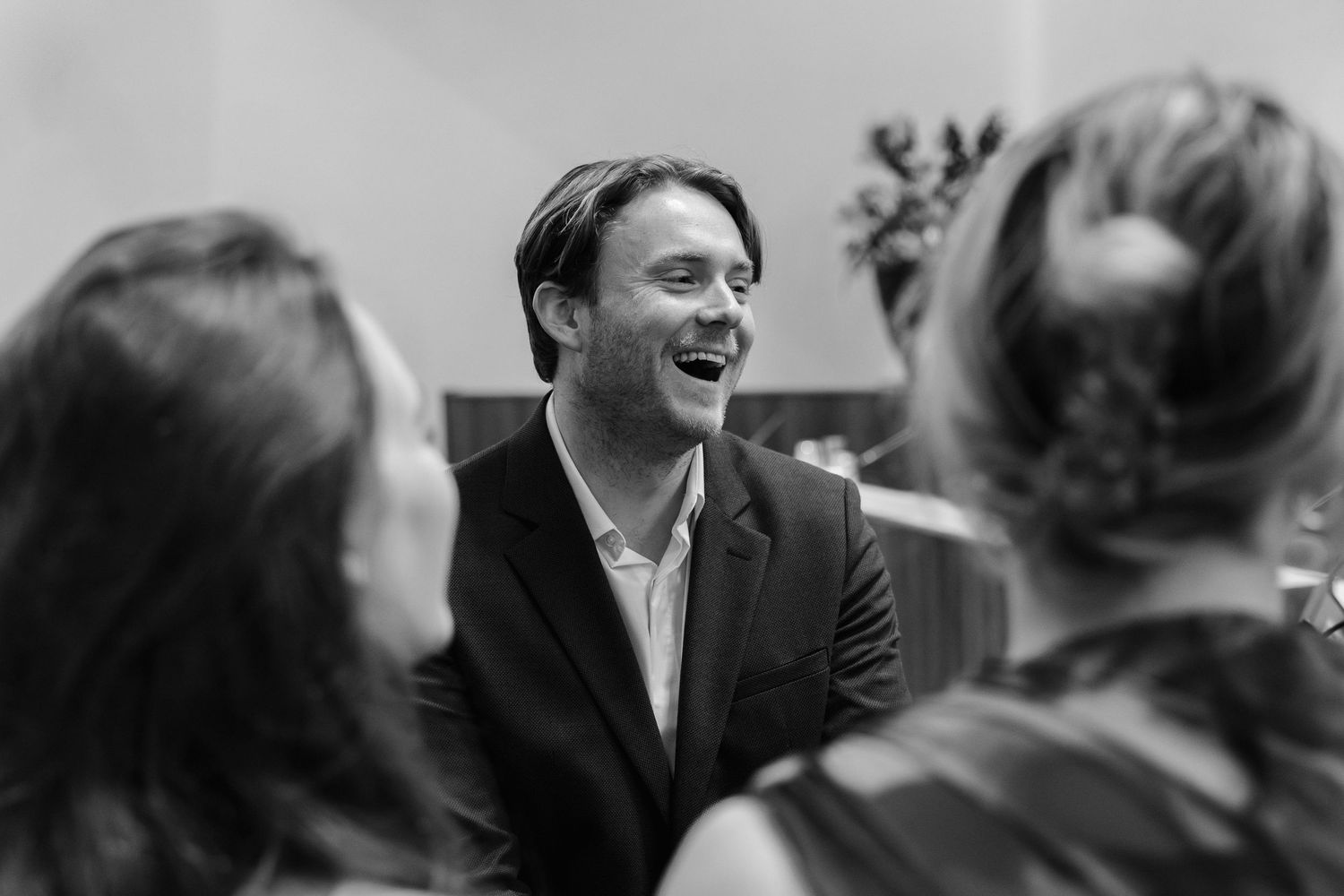 A man in a blazer laughs joyfully while socializing with guests at an indoor event, captured in black and white.
