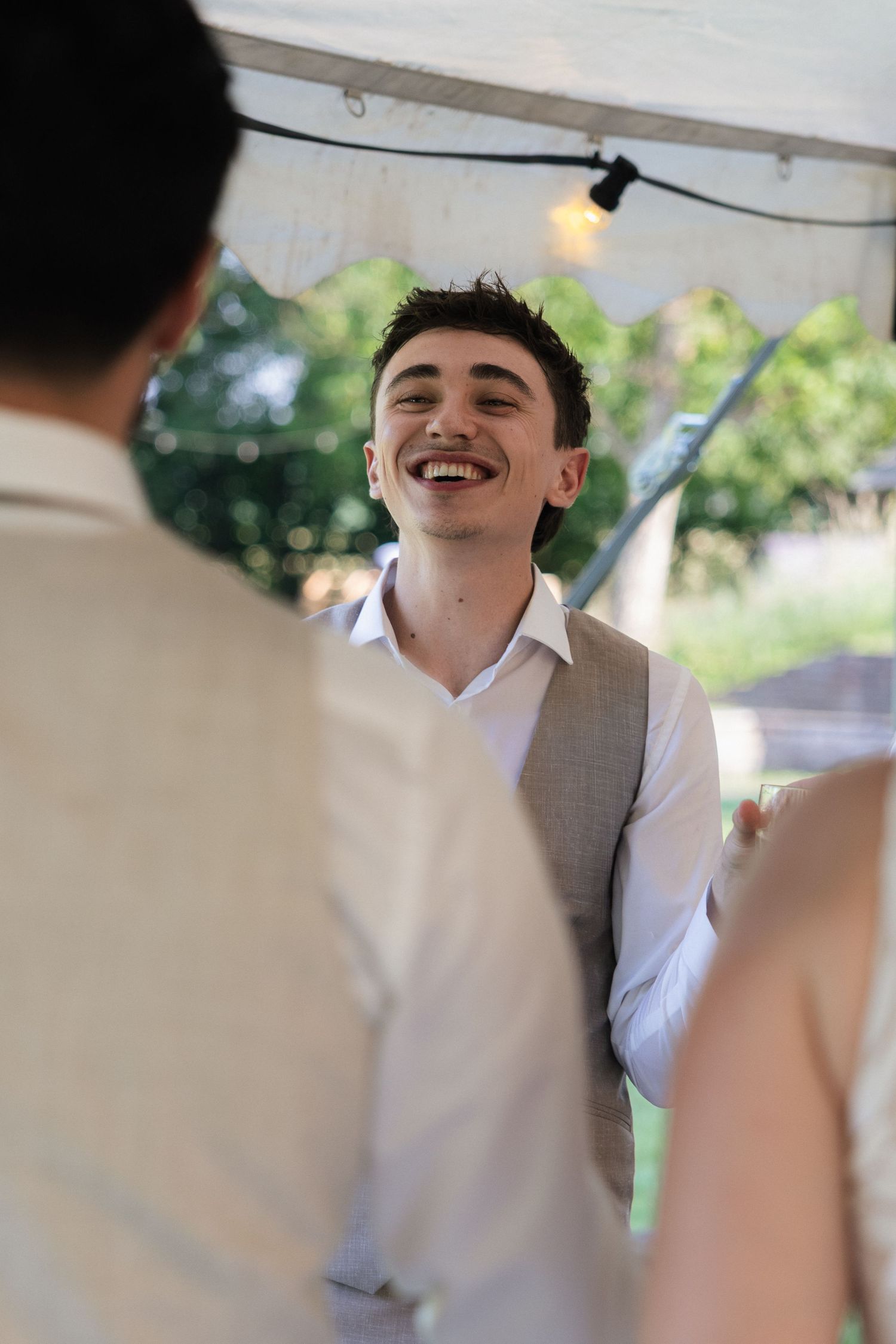 A young man in a gray vest and white shirt laughs joyfully at an outdoor wedding reception under a white tent.