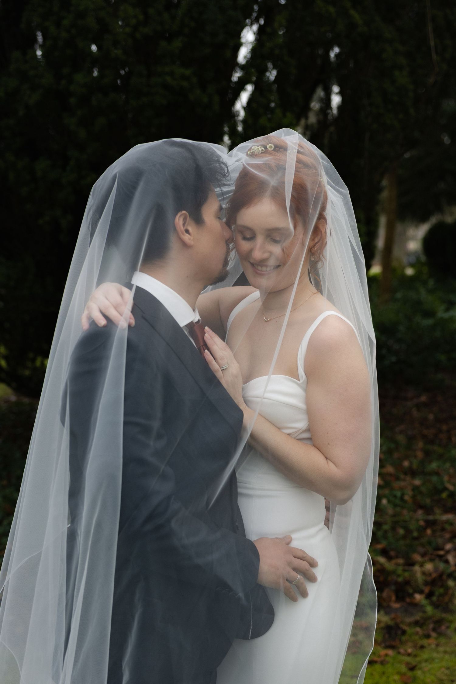 Bride and groom share romantic moment under wedding veil outdoors, bride in white dress smiling at groom in gray suit.