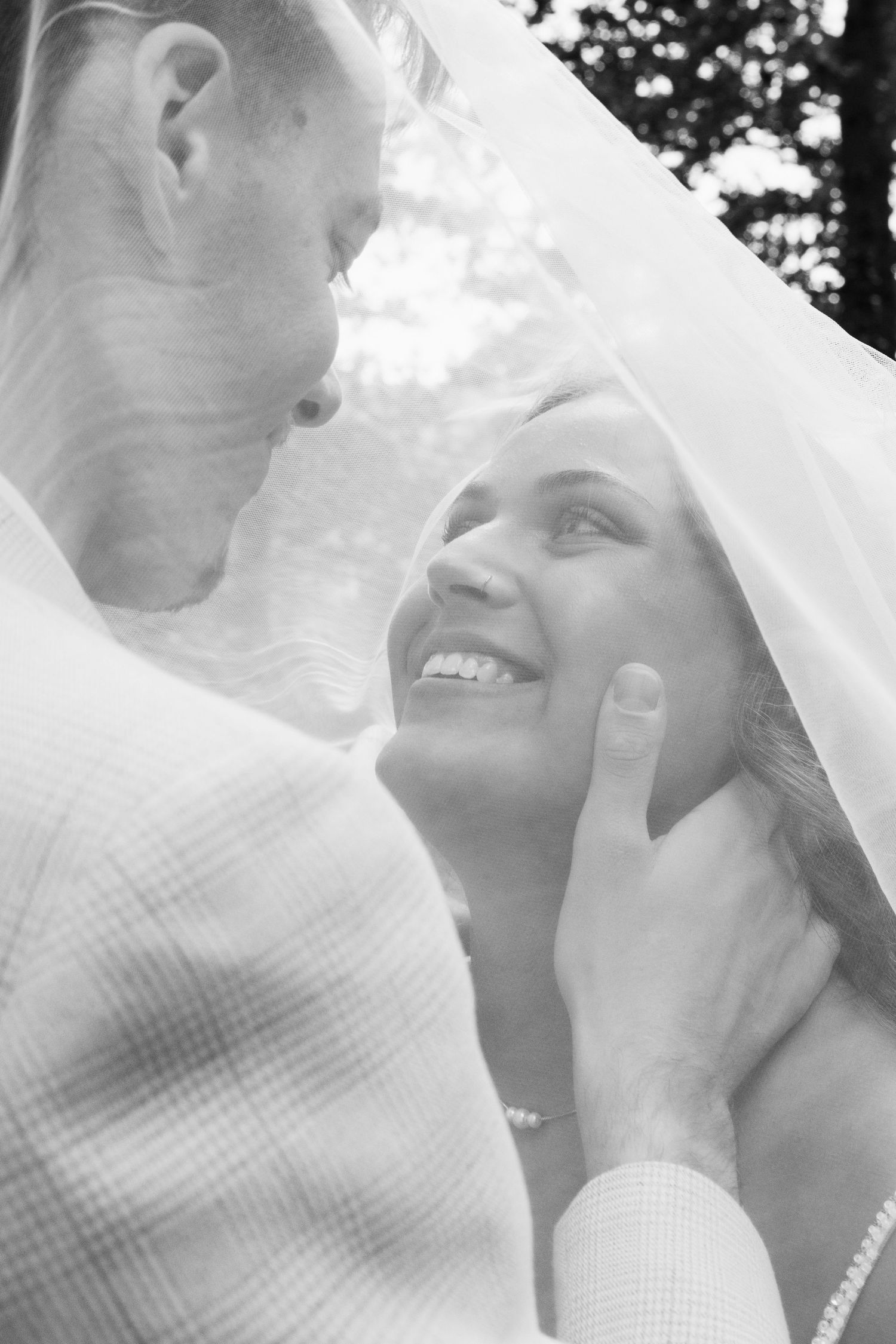 Black and white wedding photo of groom tenderly holding brides face under veil, bride smiling up at him outdoors.