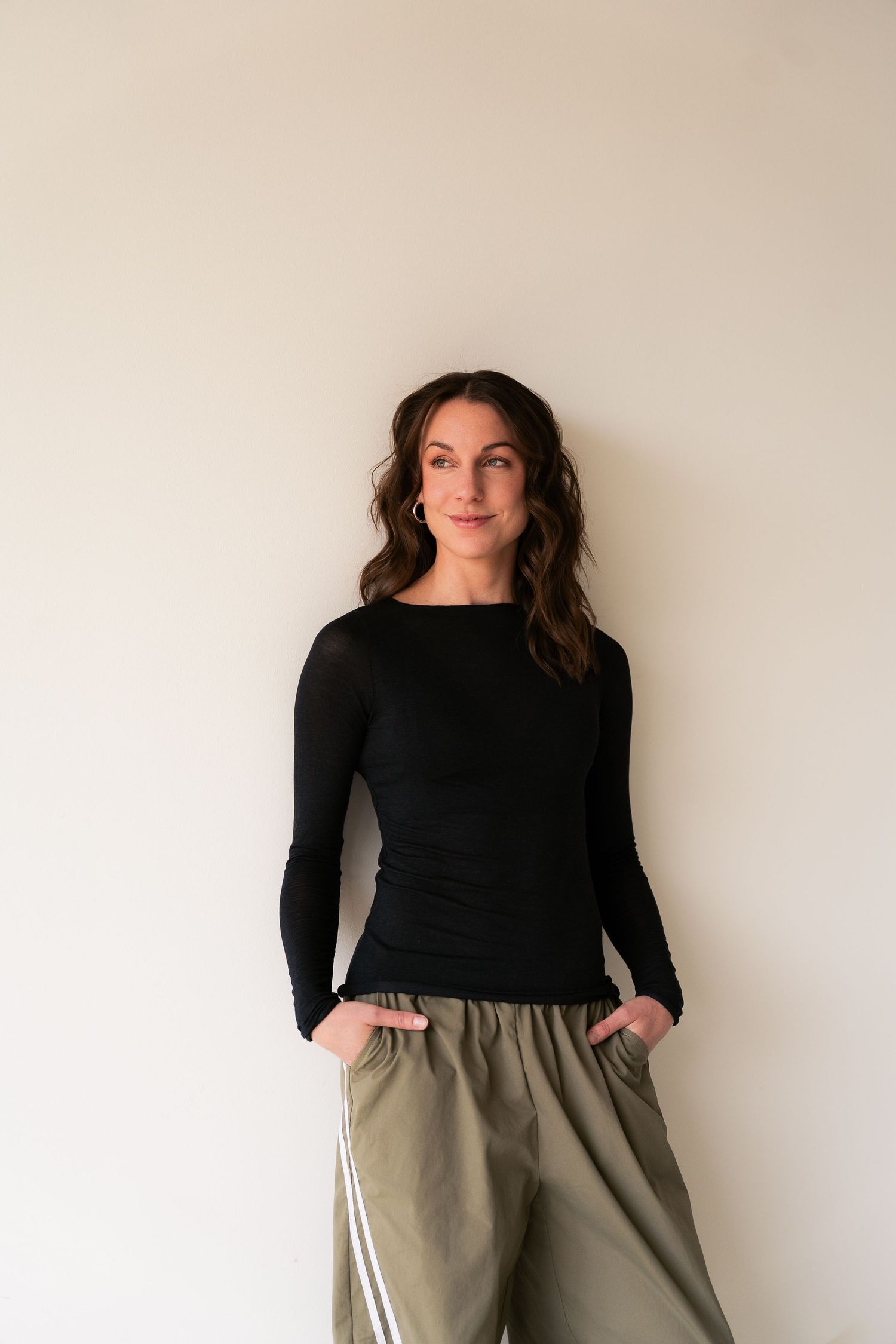 Woman in black long-sleeve top and olive green pants standing against a light wall, hands in pockets.