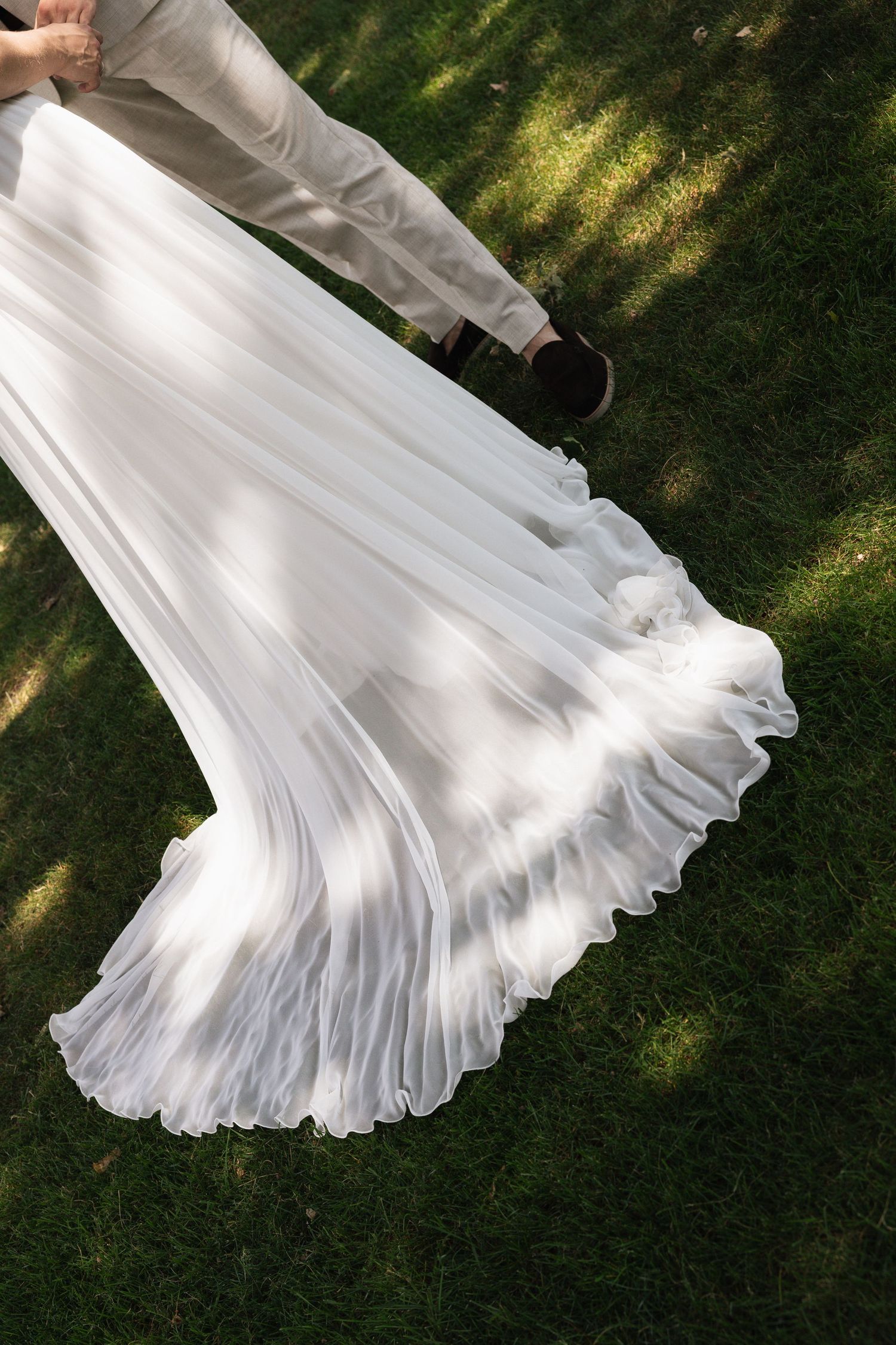 Aerial view of a bride in a flowing white wedding gown twirling on green grass beside a groom.