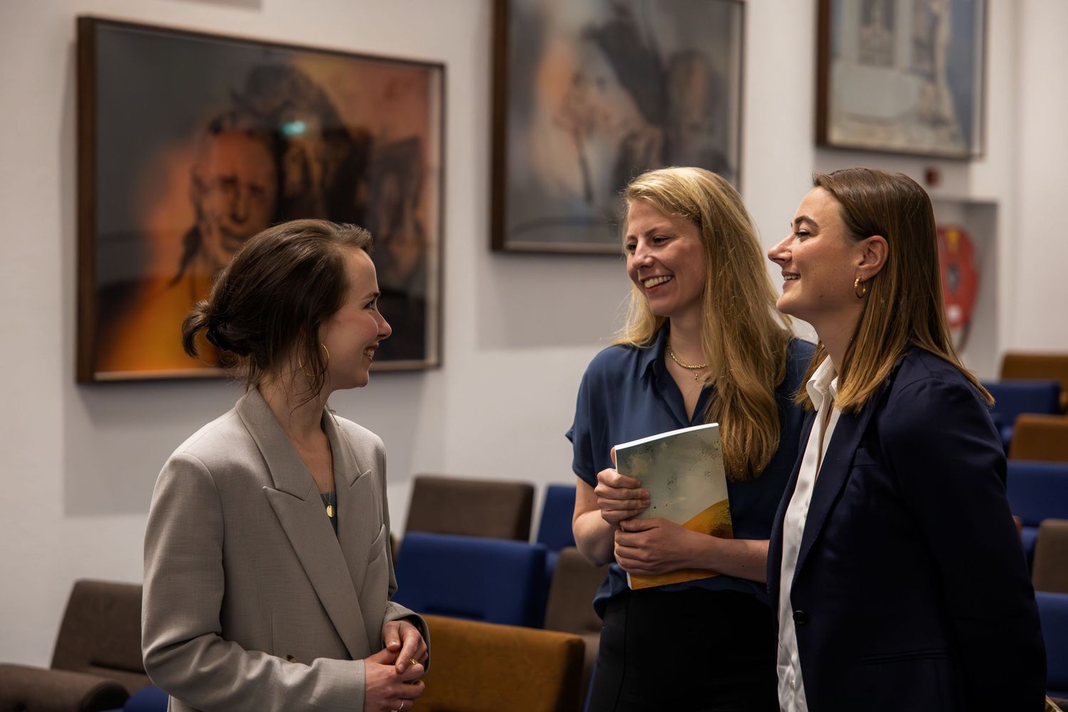 Three professional women in business attire engaged in conversation in a gallery room with artwork on the walls.