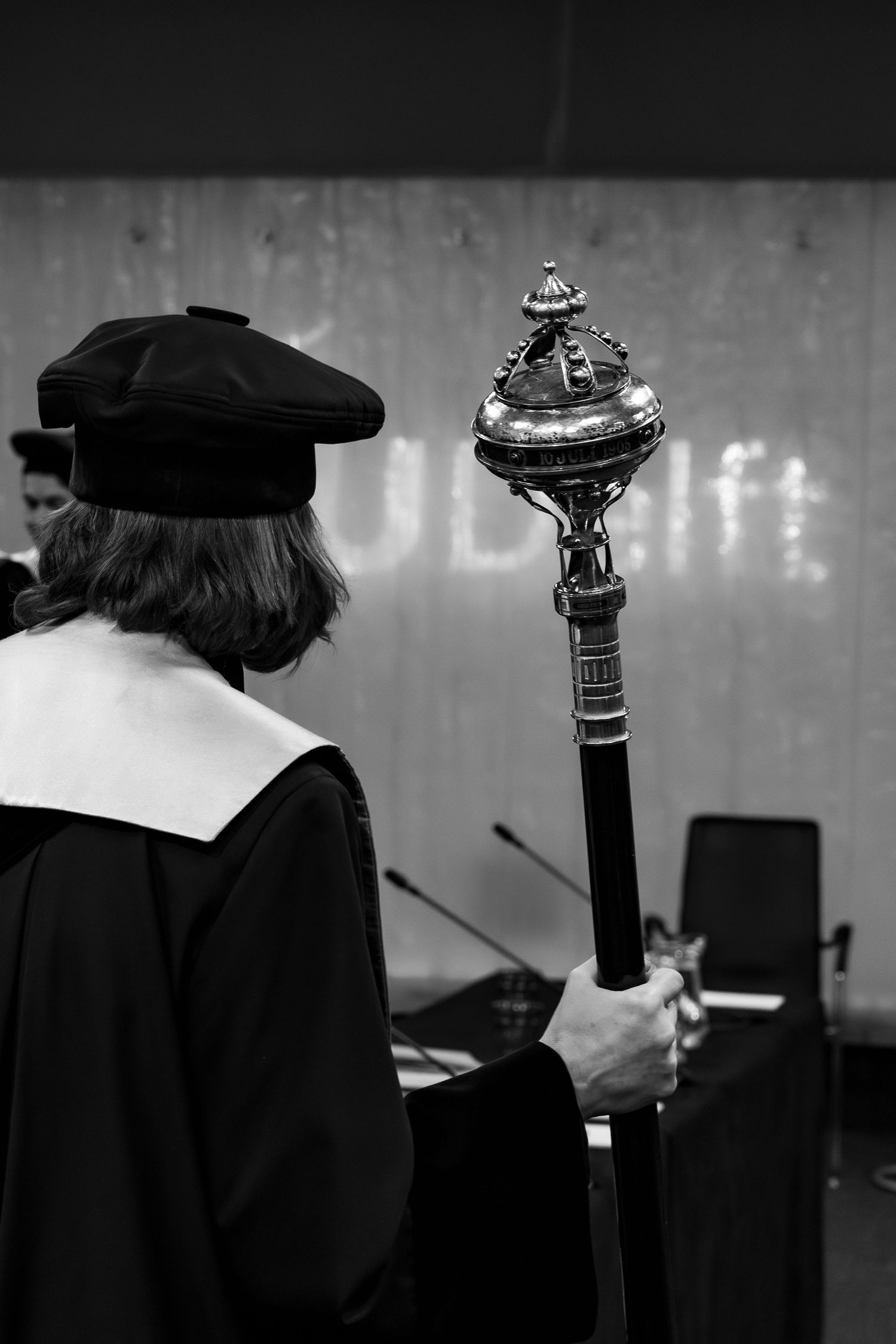 Academic dignitary in graduation robes holding ornate ceremonial mace during university commencement ceremony, black and white photo.