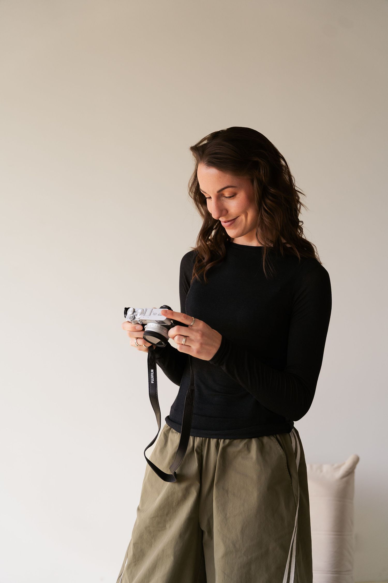 A smiling woman in a black top and olive pants holds a camera, reviewing photos in a bright studio setting.