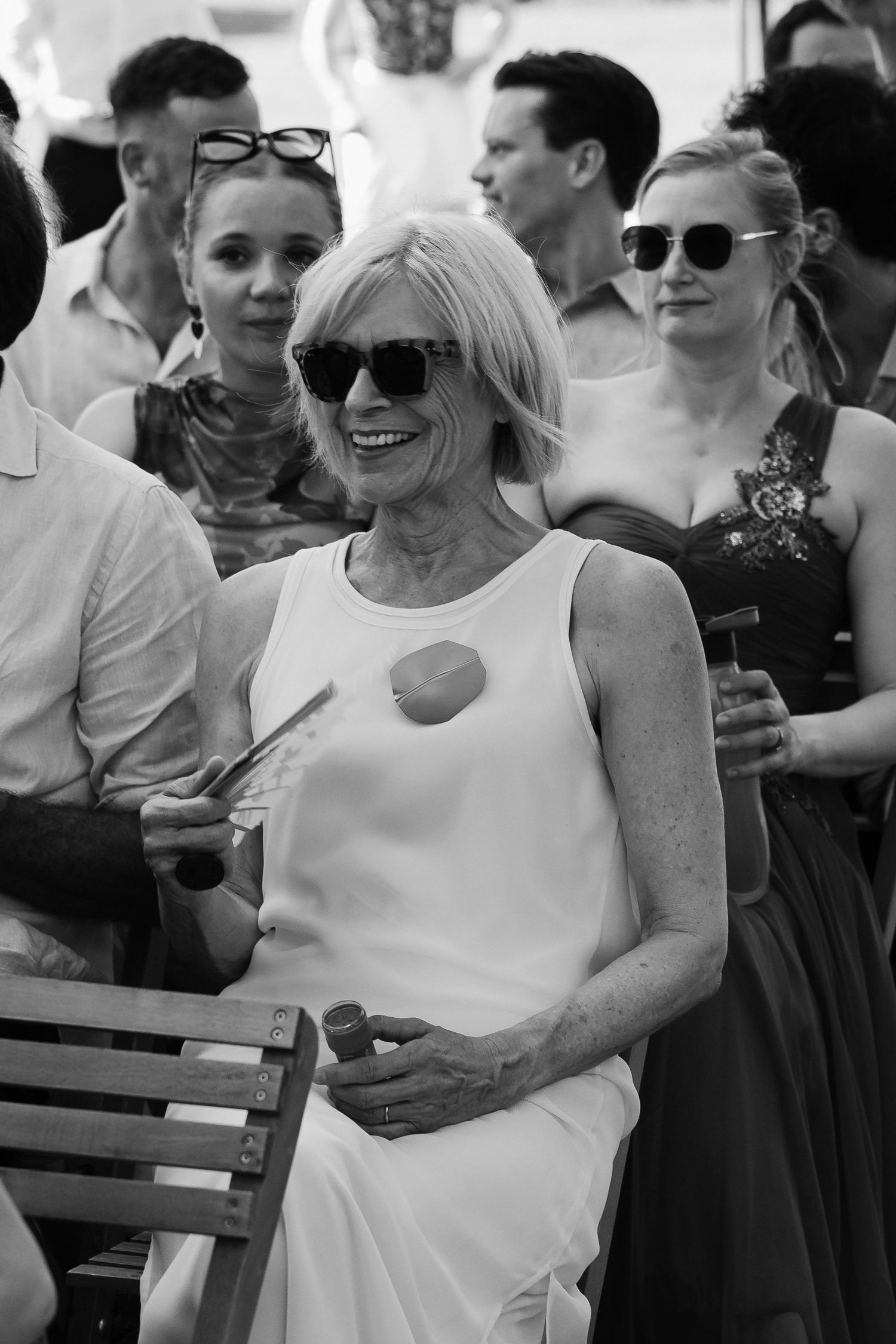 Elderly woman in white dress and sunglasses smiling at outdoor event with guests in background.