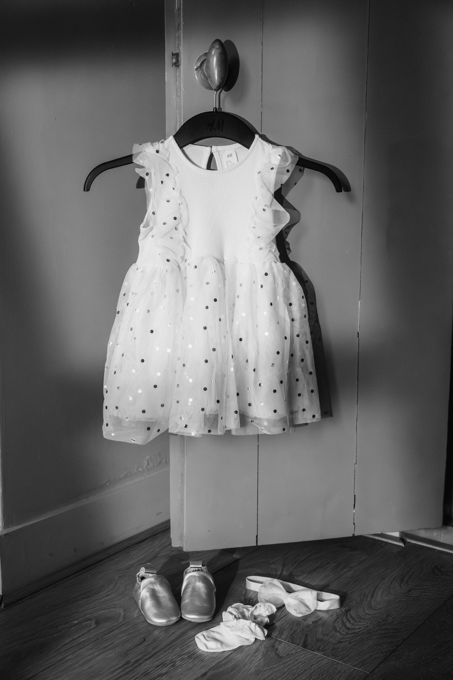 Black and white photo of a polka dot baby girl dress on hanger with tiny shoes and socks on wooden floor.