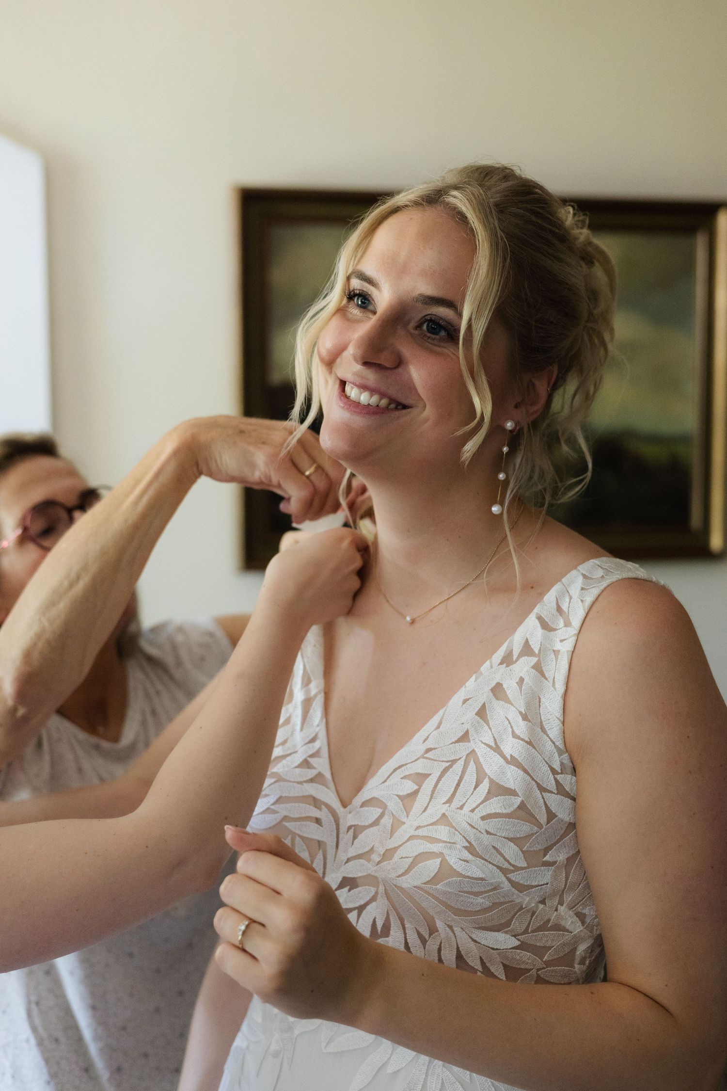 Smiling bride in lace wedding dress having earrings put on during bridal preparations.