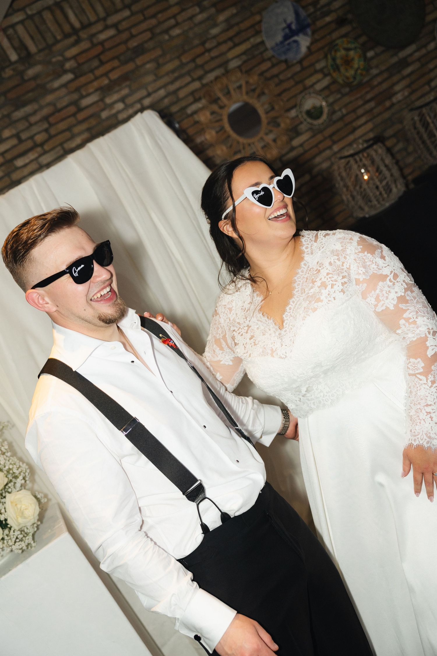 Bride in lace gown and groom in white shirt with suspenders wearing fun sunglasses at wedding reception.