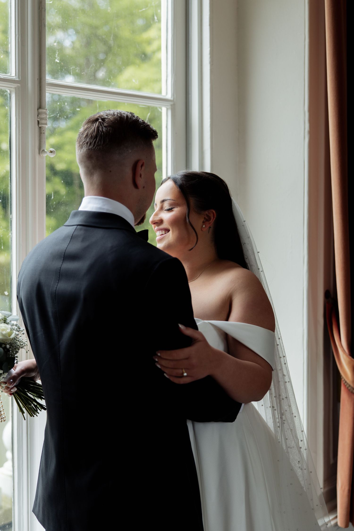 Bride and groom share a romantic moment by a sunlit window, with bride in white gown and veil smiling at her partner.