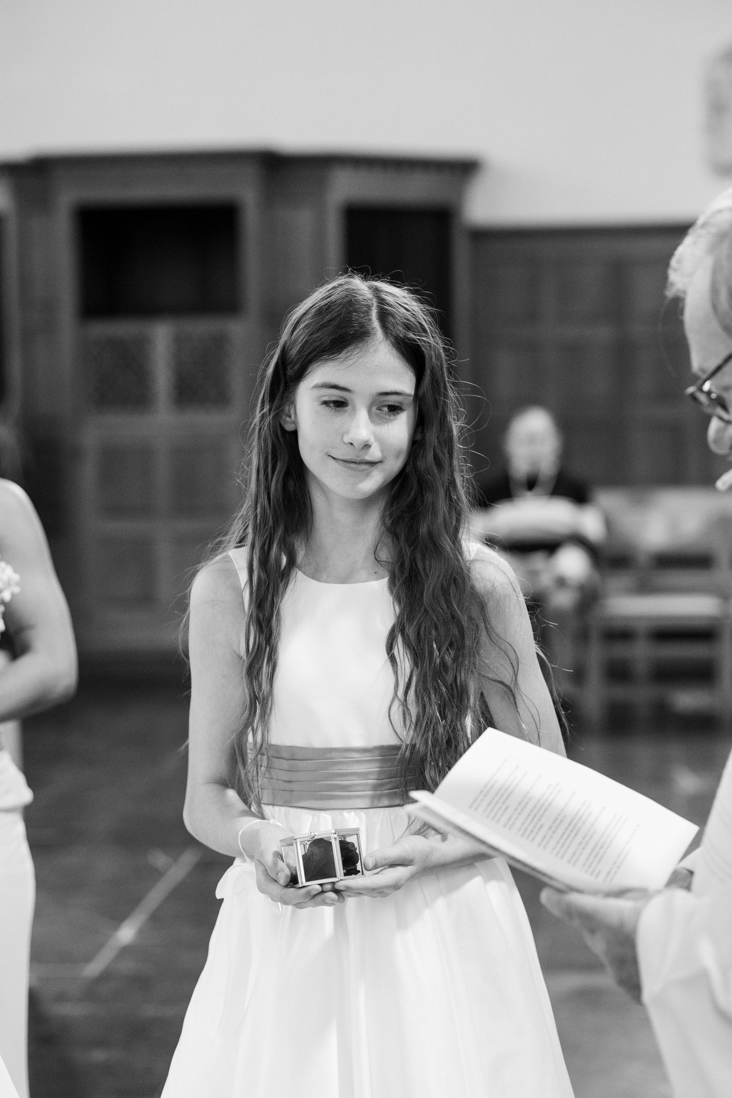 Young girl in white dress with sash holds rings at wedding ceremony, black and white photo.