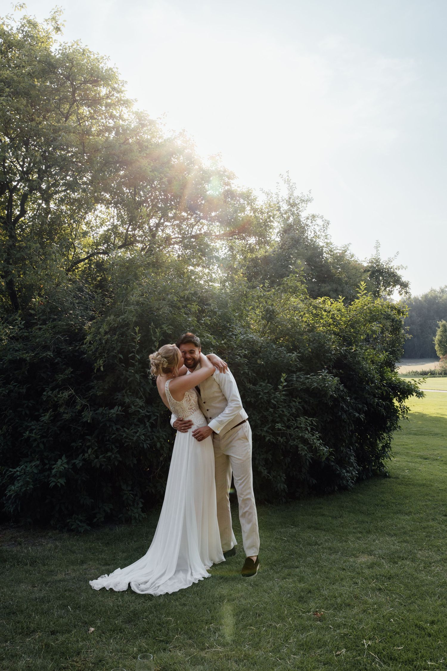 Bride and groom embracing in golden sunlight on green lawn with lush trees, romantic outdoor wedding portrait.