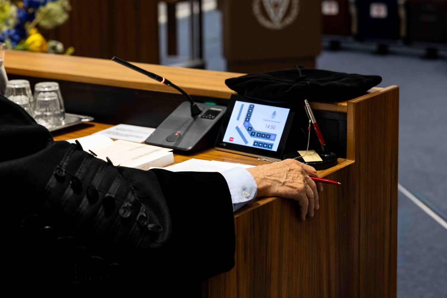 Close-up of a speaker at a podium with a tablet, microphone, and documents in a formal conference room setting.